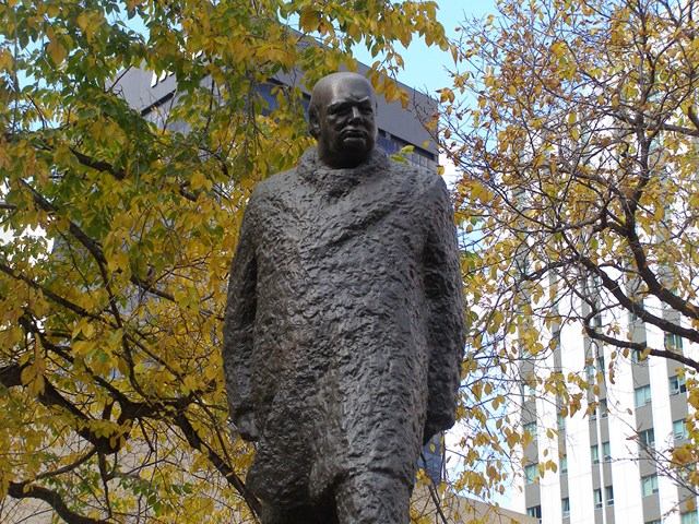 Bronze statue of Winston Churchill surrounded by autumn trees with city buildings in the background.
