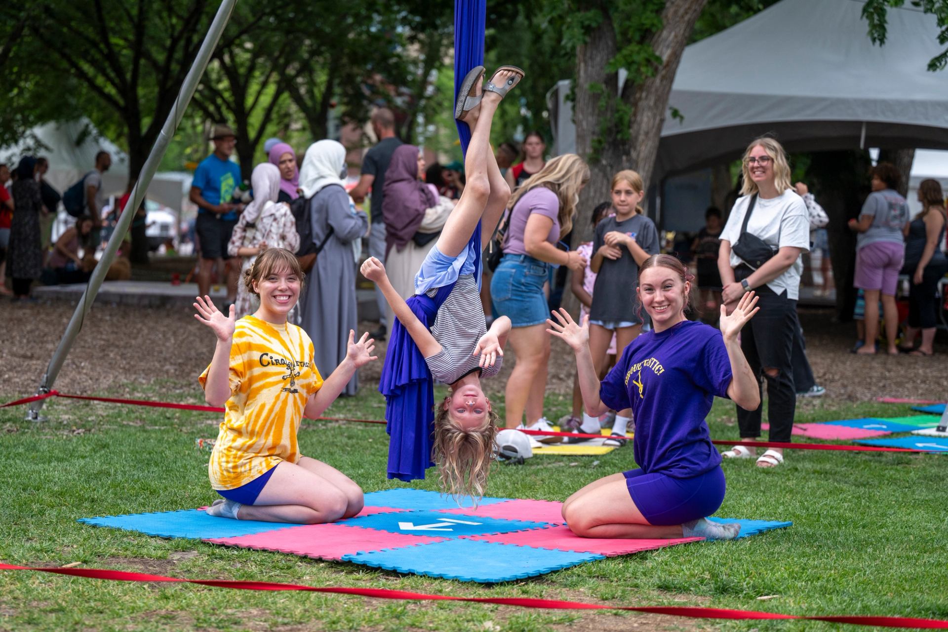 Outdoor festival scene with child hanging from aerial silks between two kneeling performers.