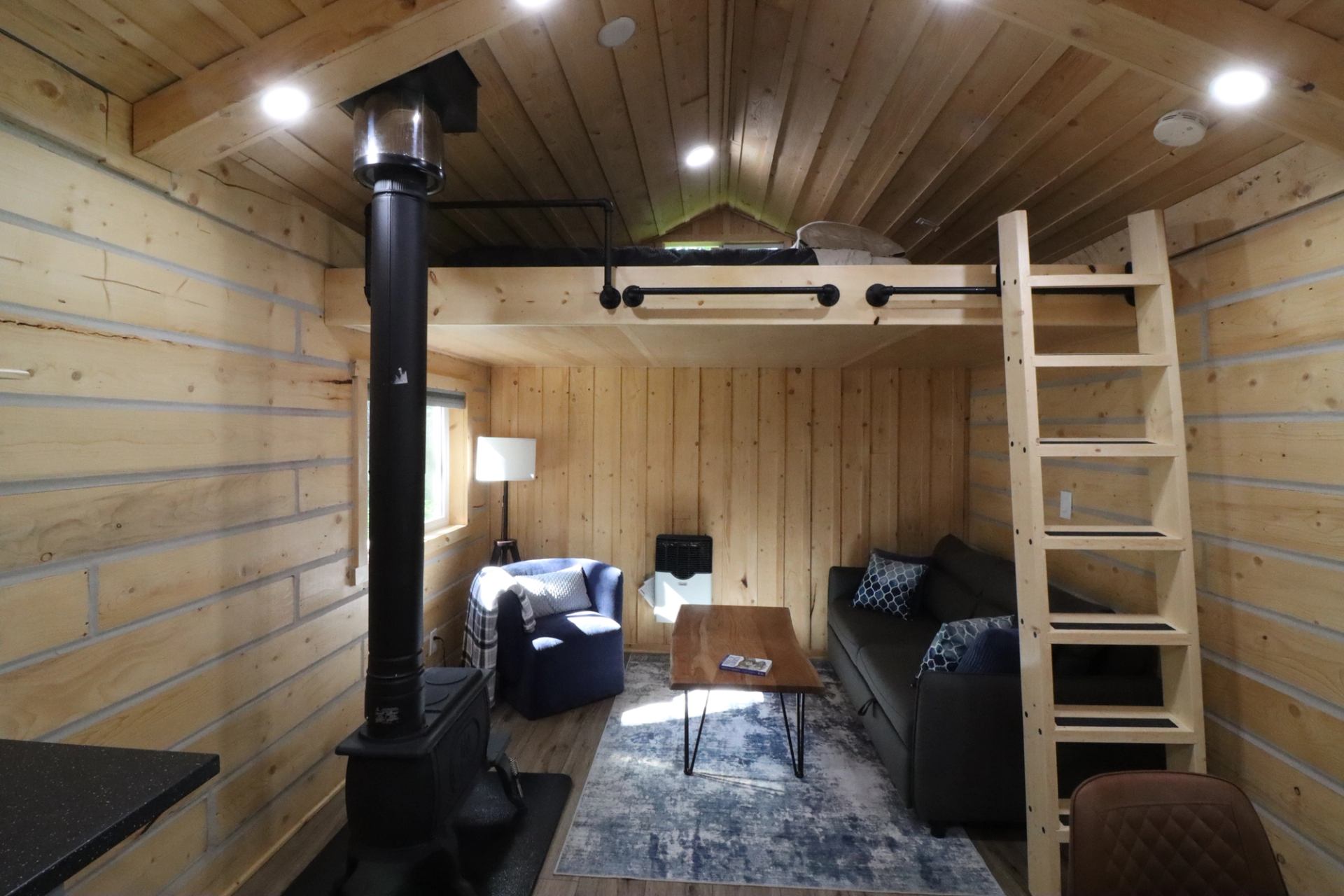 Cozy cabin interior with loft, stove, sofa, armchair, coffee table, and warm wood finishes.