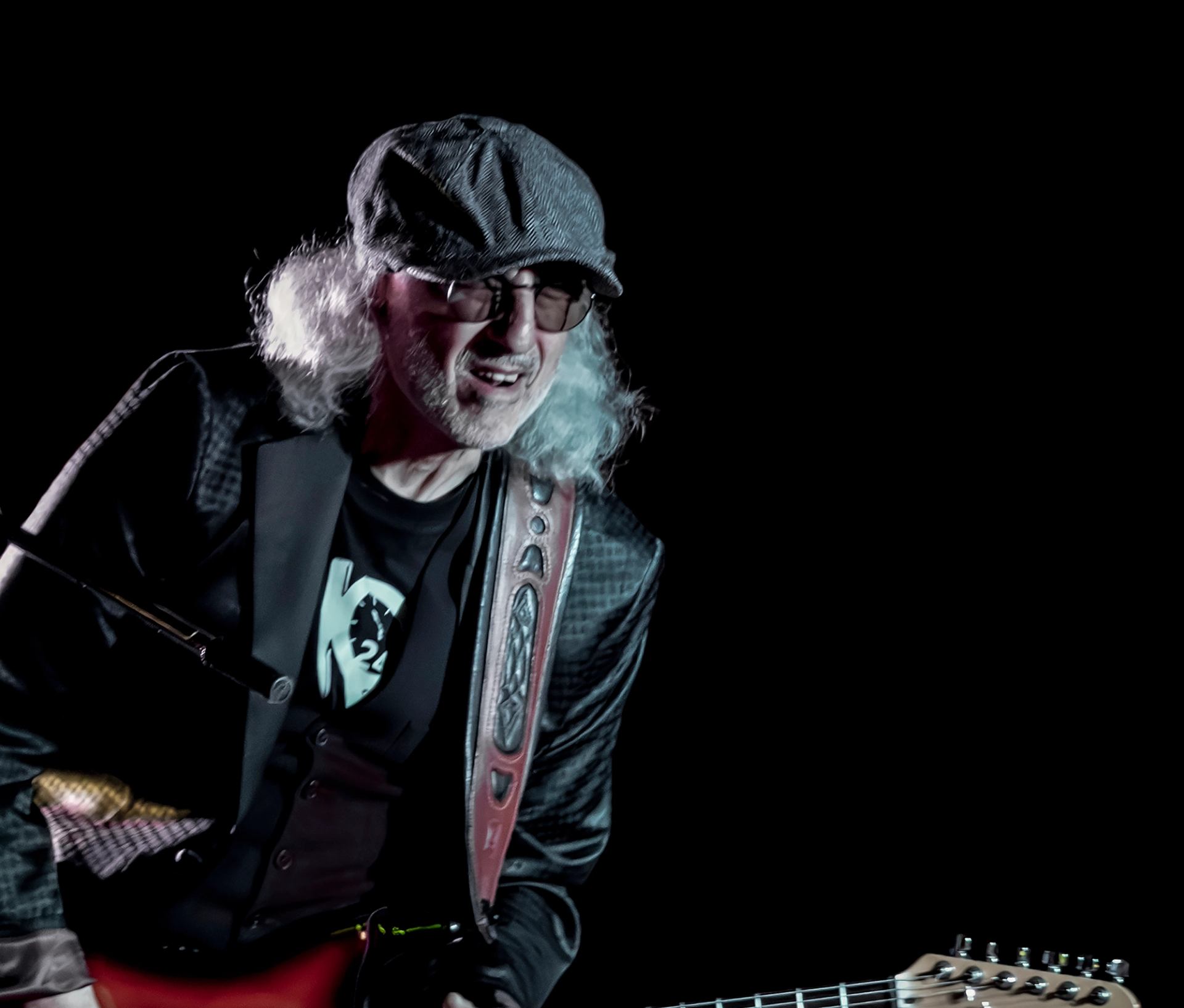 Musician playing an electric guitar on a dark stage, wearing a hat and jacket under dramatic lighting.