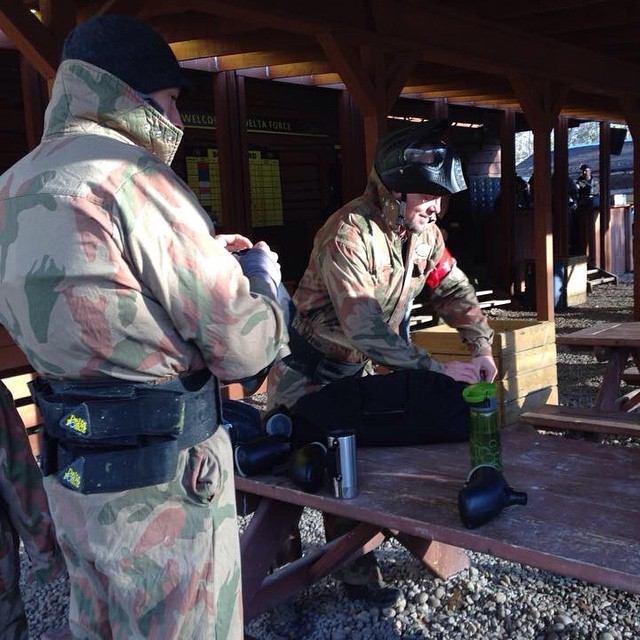 Paintball players in camouflage gear adjust equipment on picnic table under wooden shelter.