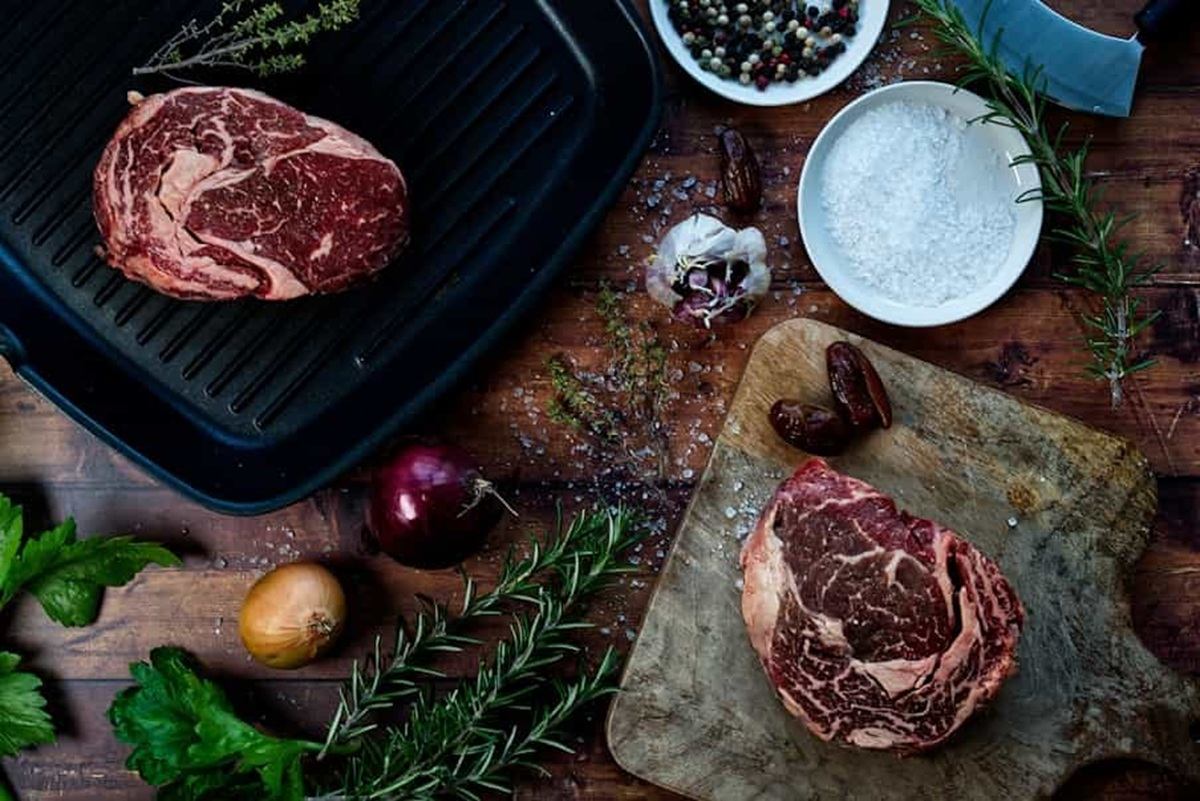 Raw ribeye steaks on grill pan and cutting board with herbs, onions and seasonings.
