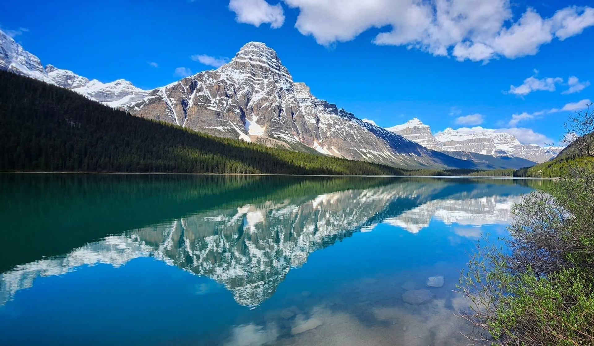 Snow-capped mountain and forest reflected in a calm turquoise lake under blue sky.