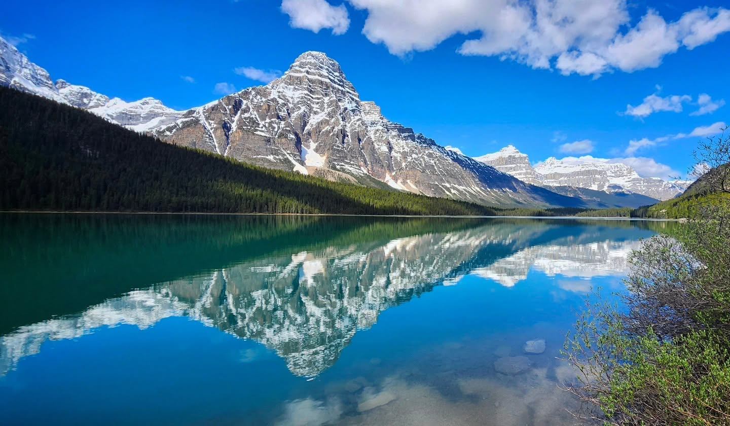 Snow-capped mountain and forest reflected in a calm turquoise lake under blue sky.