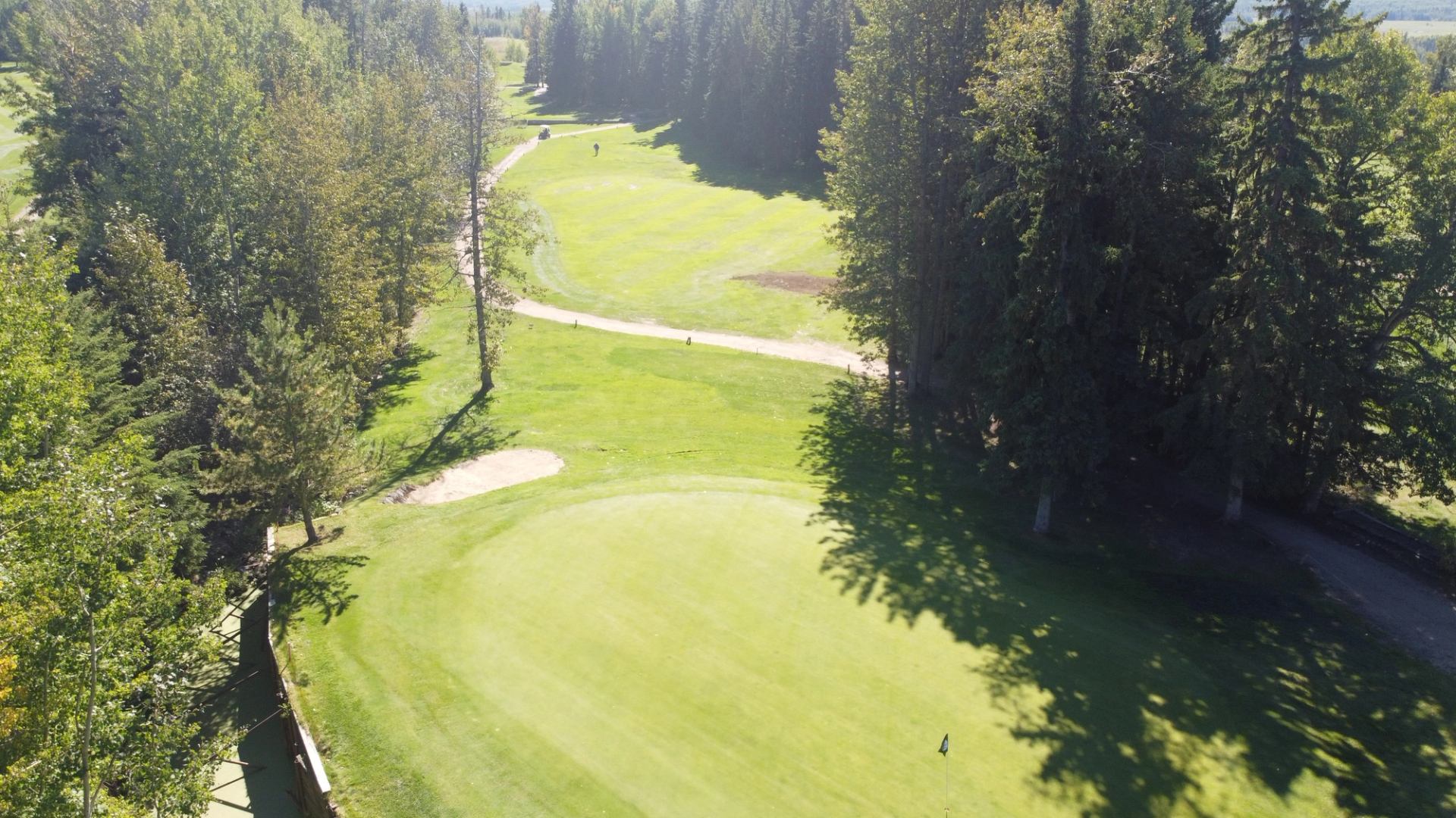 Aerial view of a green golf course with fairways, trees, a sand trap, and a flagstick.