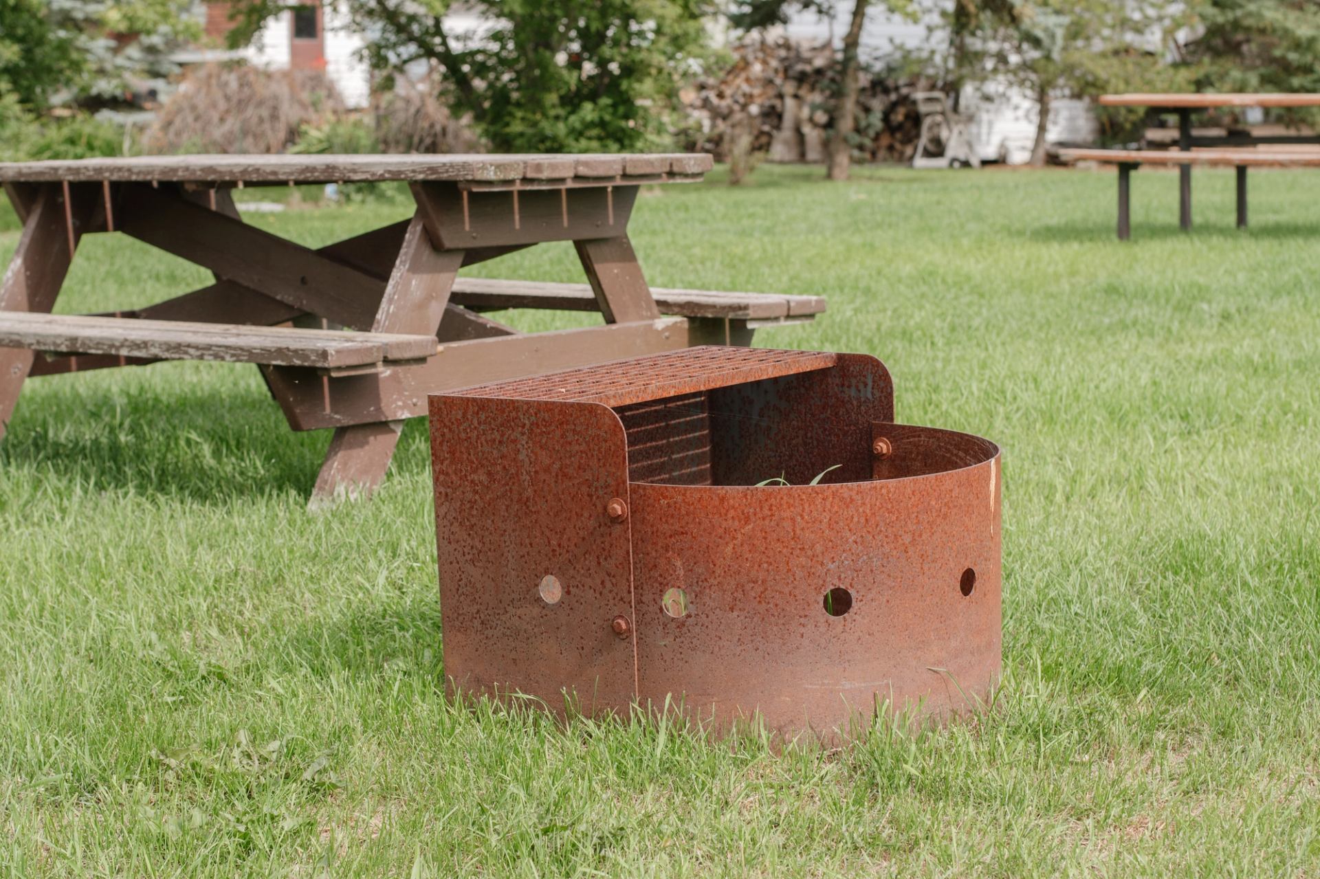 Rusty metal fire pit on grass near wooden picnic tables in a park.