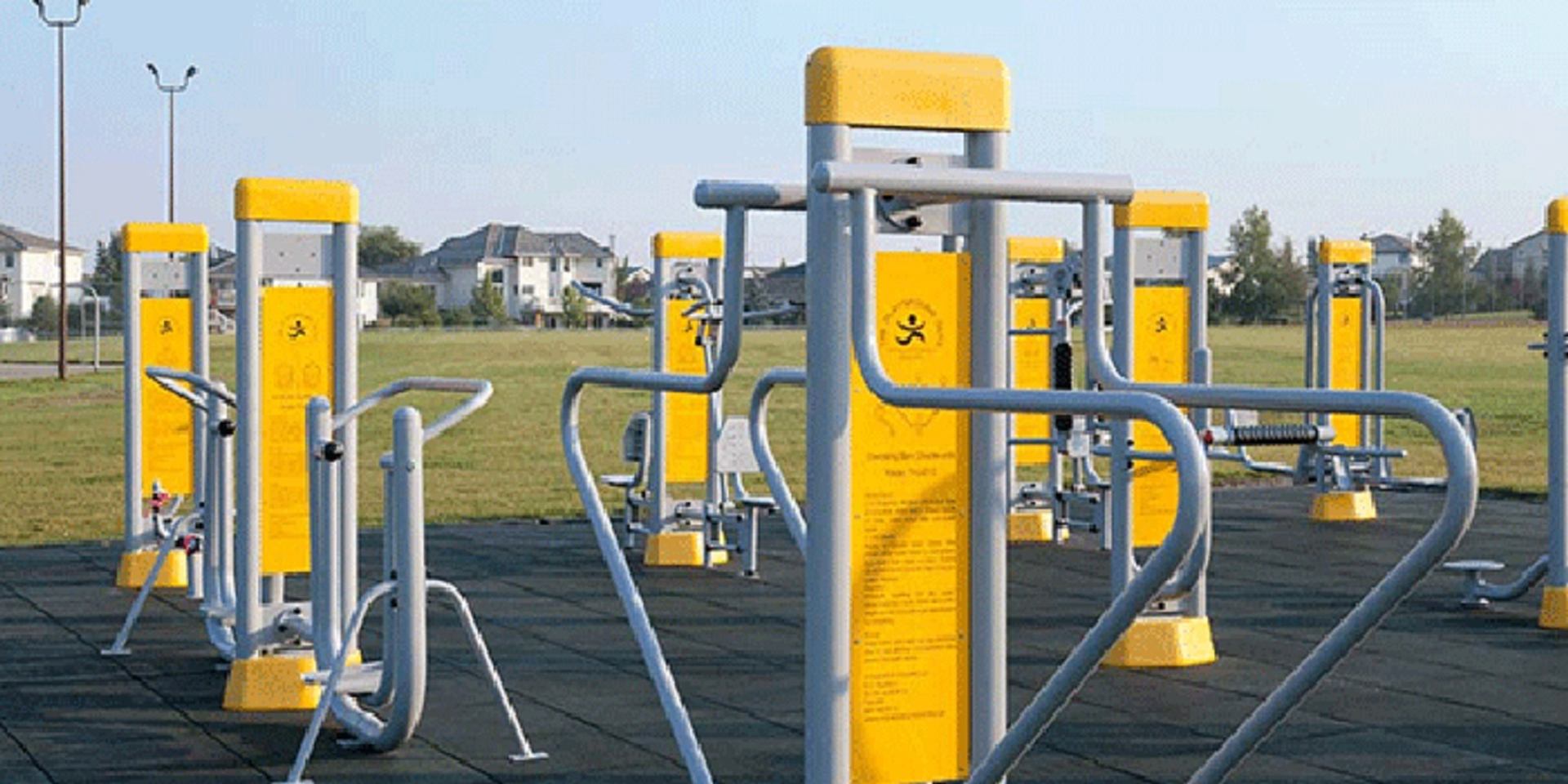 Outdoor fitness equipment with yellow panels on rubber flooring in a grassy park near houses.