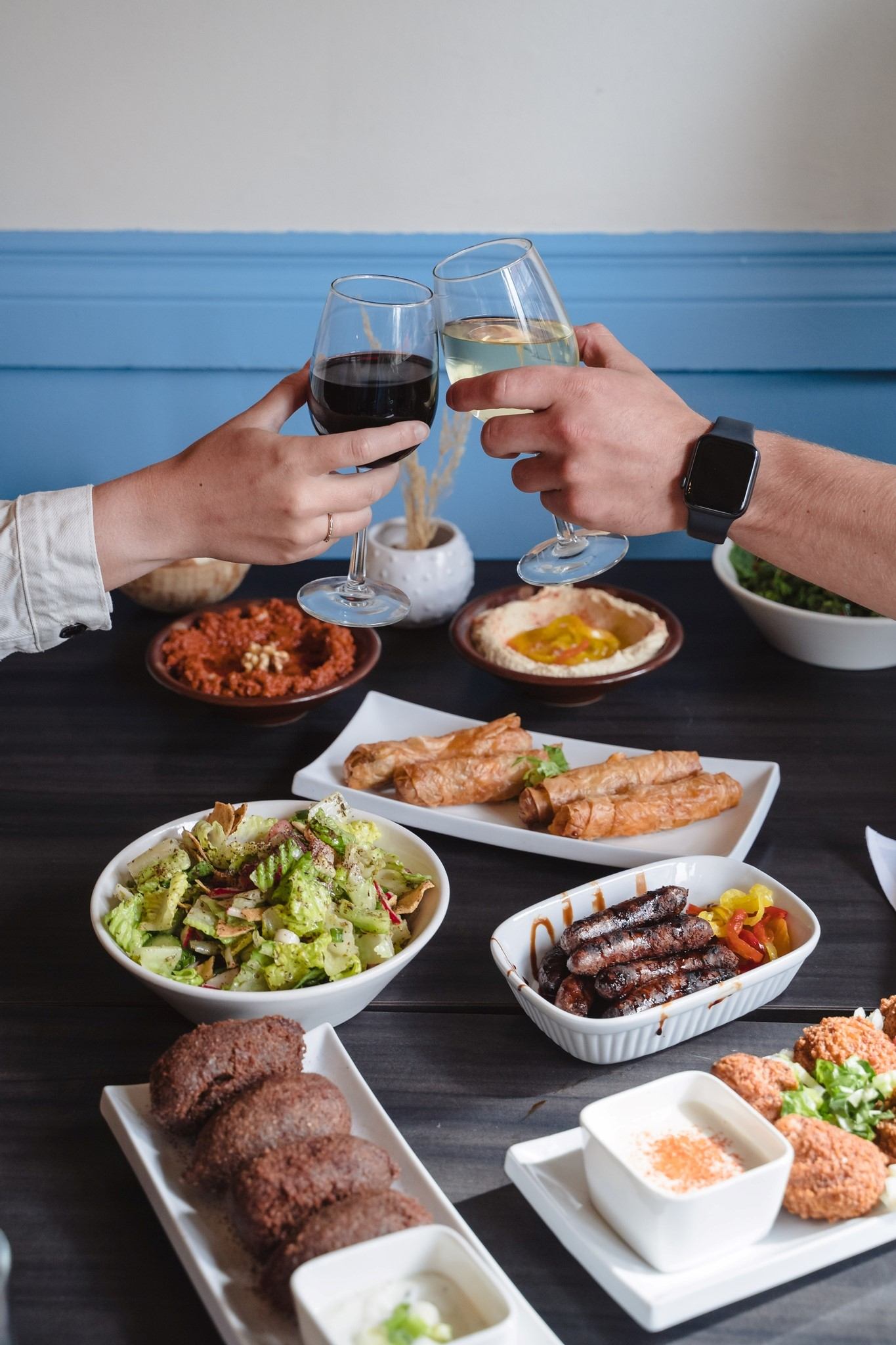 Two hands clinking wine glasses over a table of Mediterranean dishes at Aida’s Bistro.