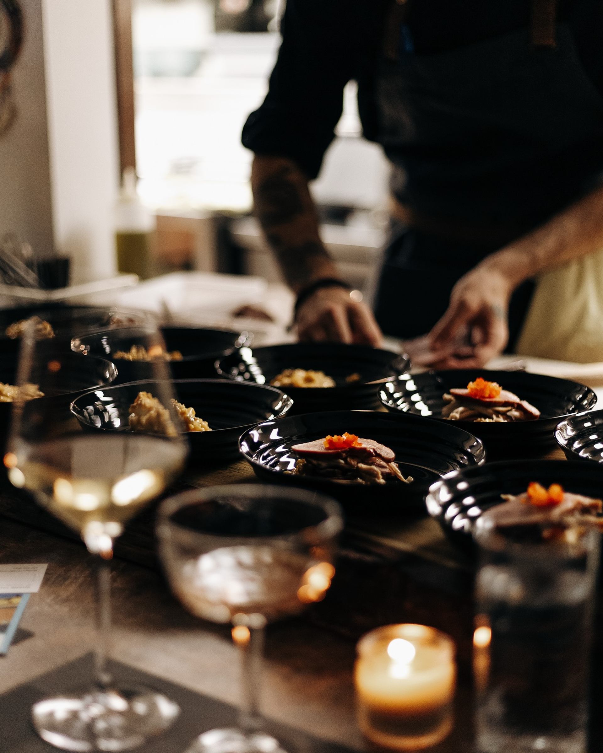 Chef plates multiple black dishes of food beside wine glasses and a lit candle.