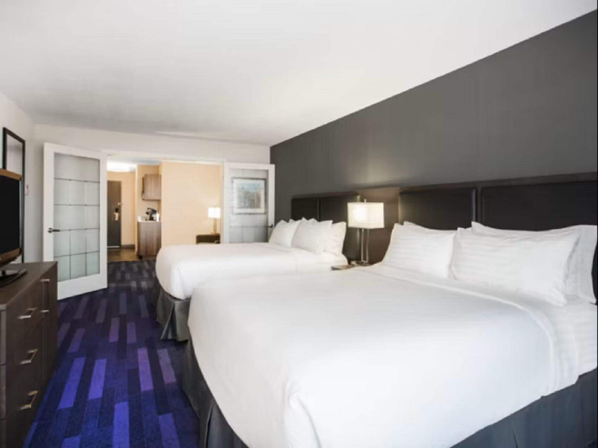 Hotel room with two beds, accent wall, lamps, and doorway to suite area.