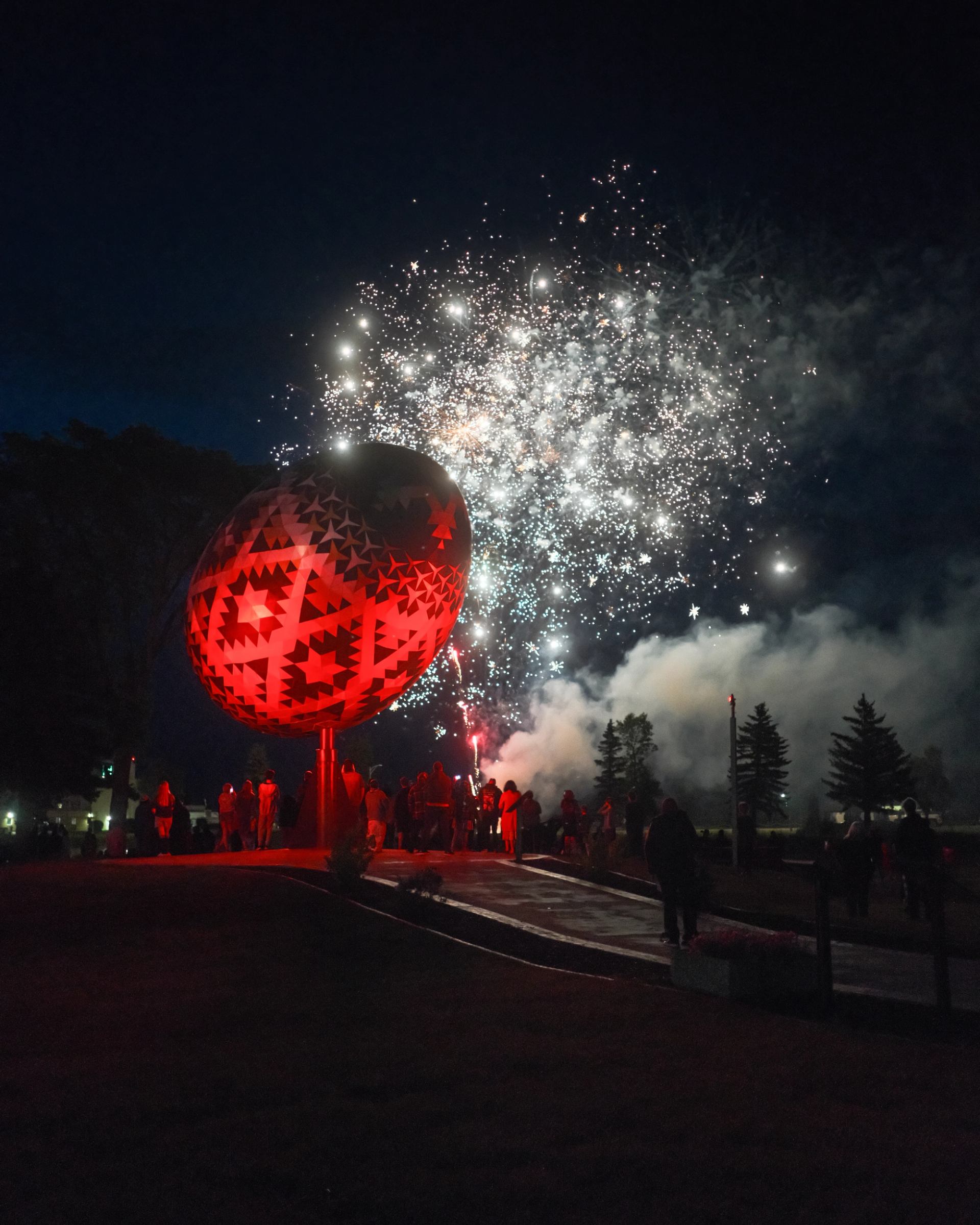 Red illuminated sphere and fireworks light up the night with silhouetted onlookers.
