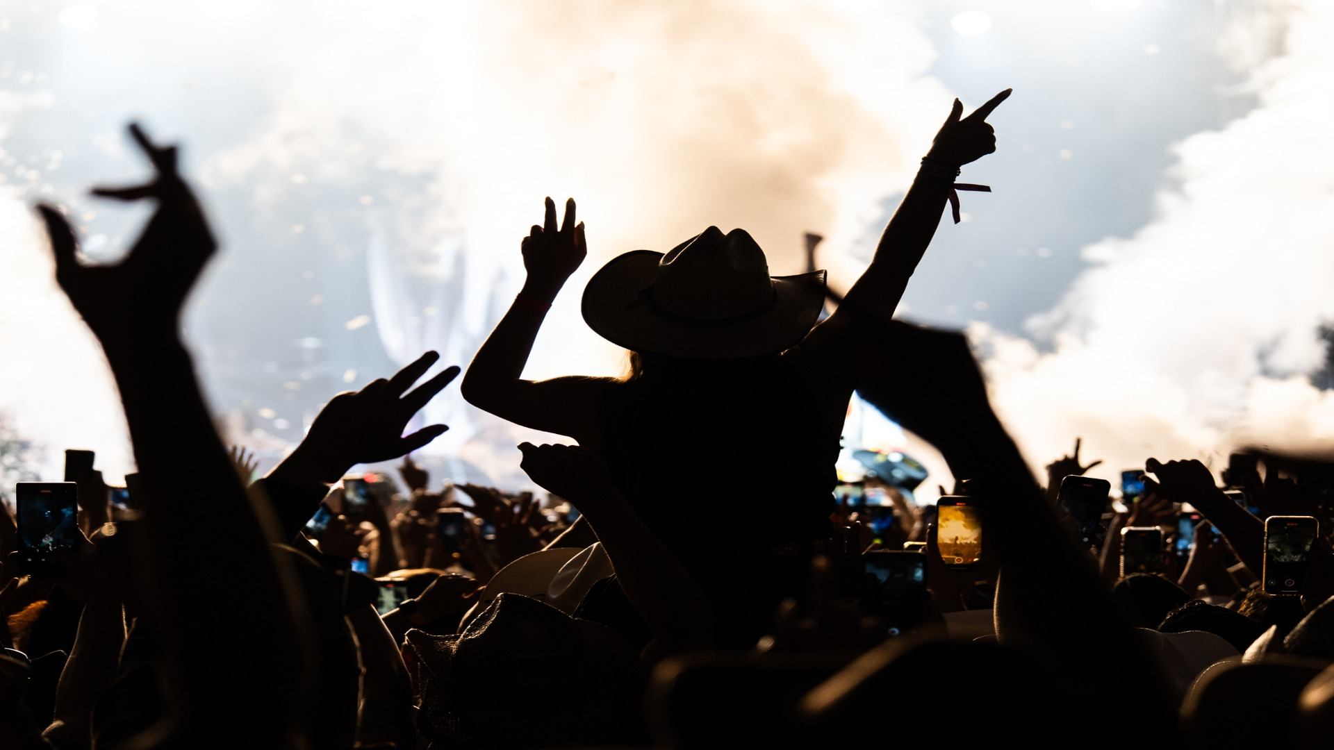 Silhouetted crowd raising hands as bright lights and smoke illuminate the festival stage.