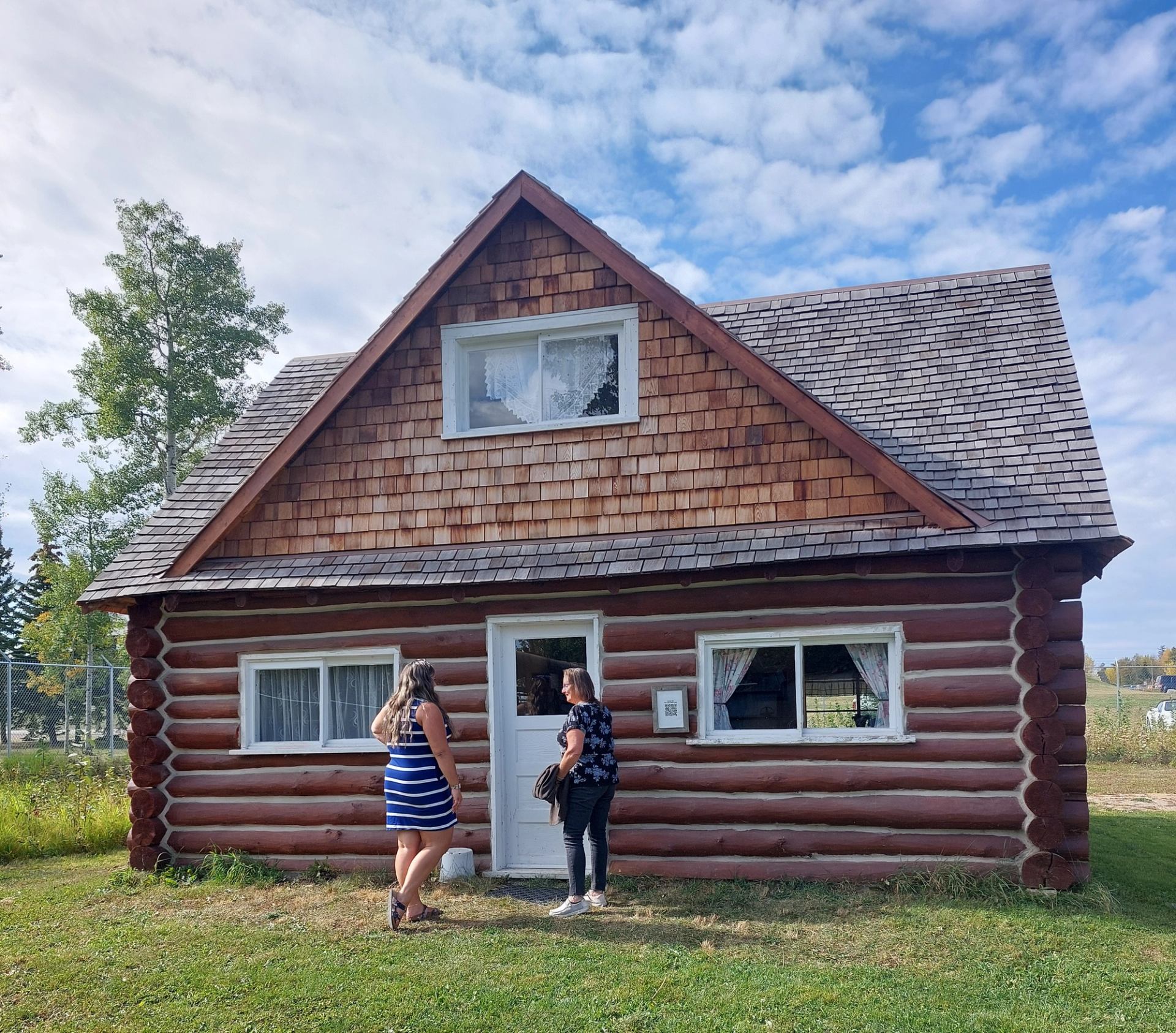 Two women stand talking in front of a rustic log cabin.