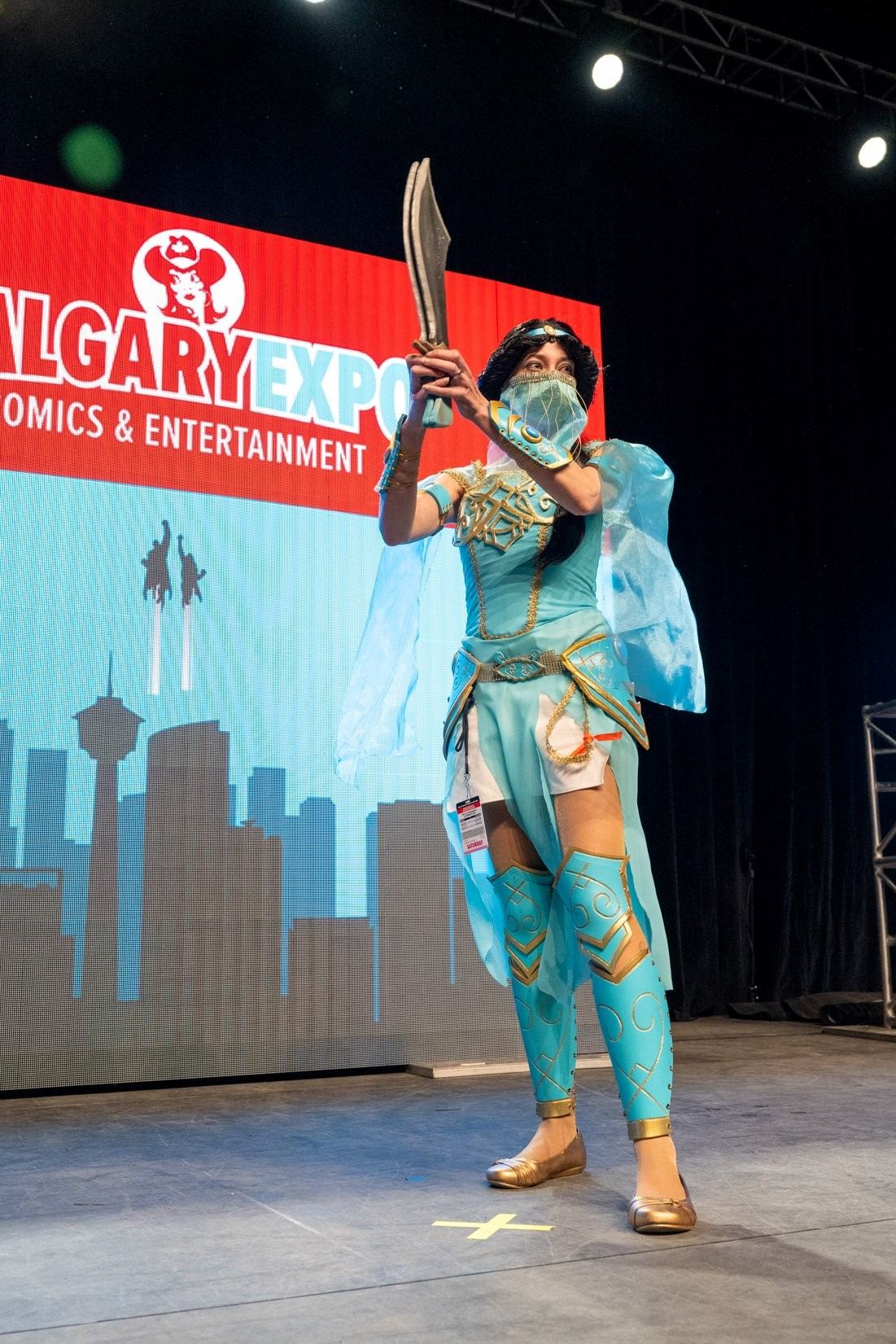 Cosplayer in a turquoise fantasy costume holding curved blades on a stage with Calgary Expo branding behind.