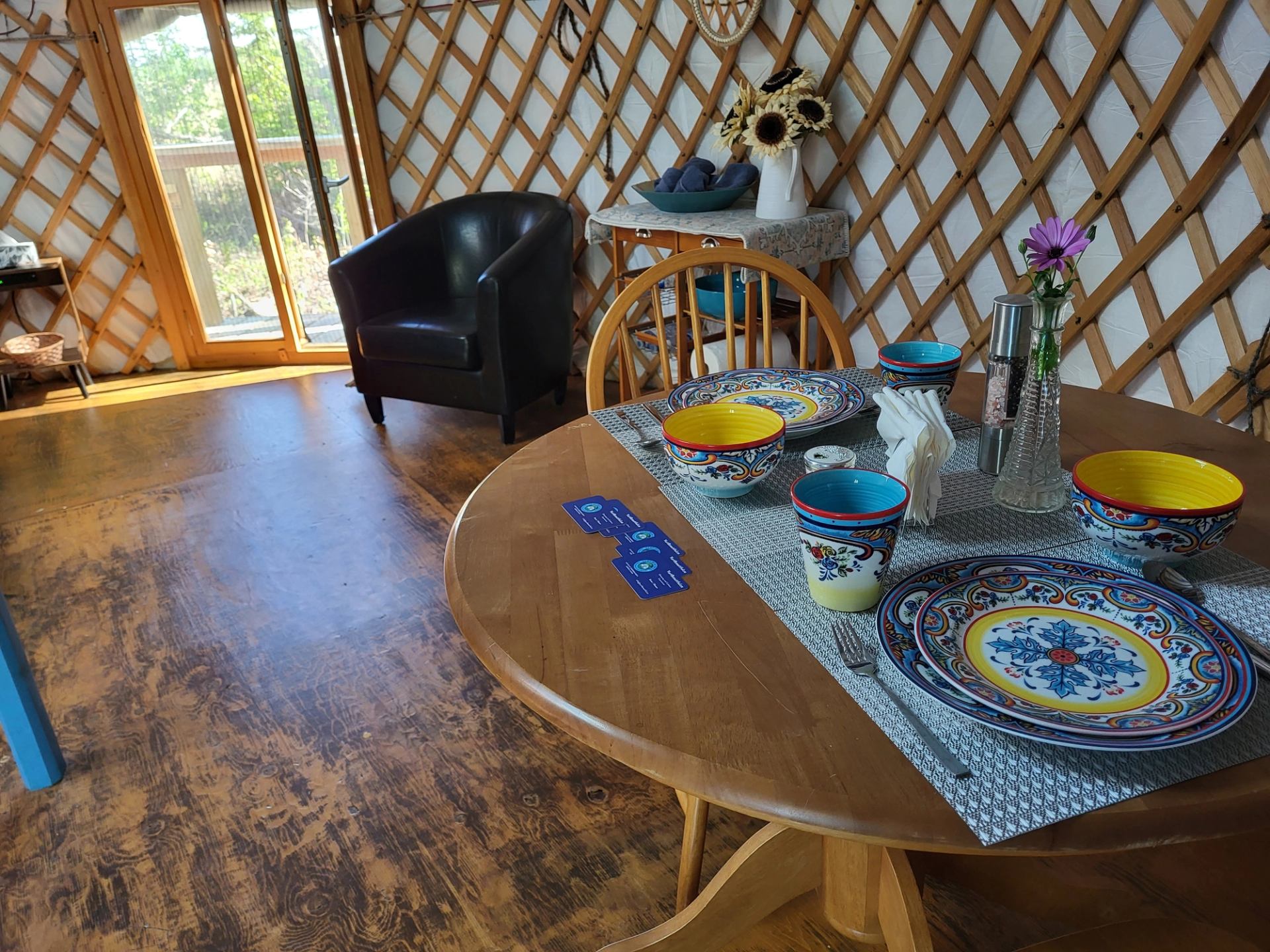 The interior of a yurt with a dining table set with colorful dishes, an armchair by a window, and sunflowers on a table in the background.