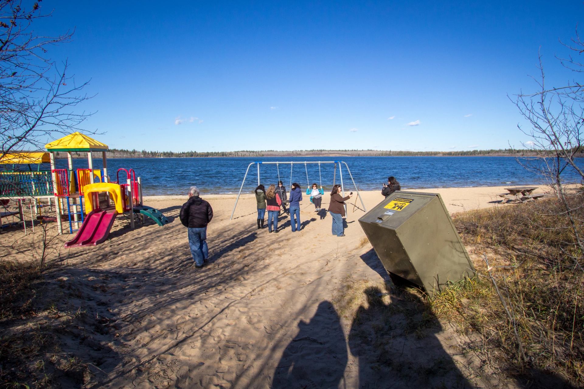 People walking on a sandy beach near a playground by a lake under a clear blue sky.