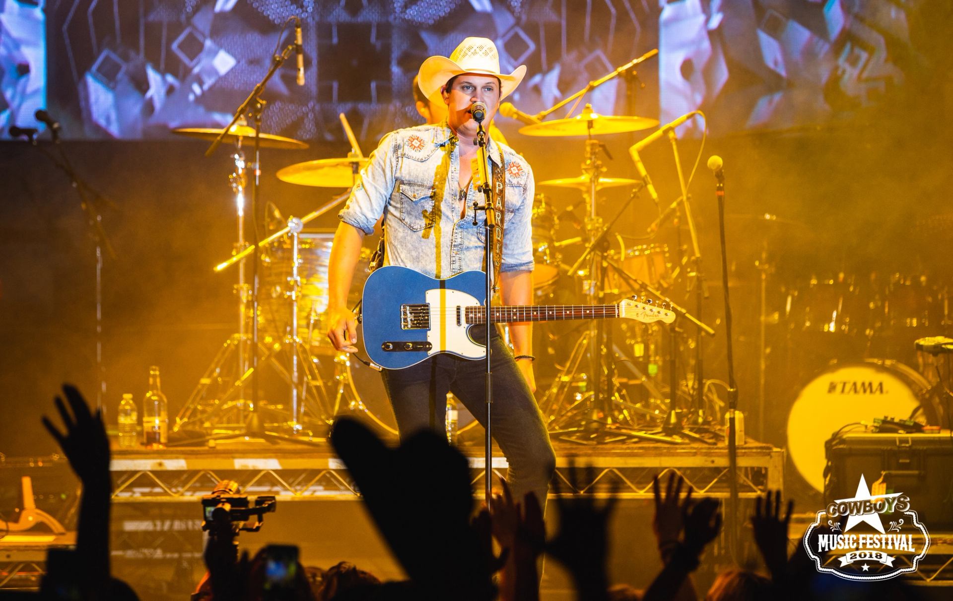 Jon Pardi playing guitar on a brightly lit stage with cheering crowd at Cowboys Music Festival.