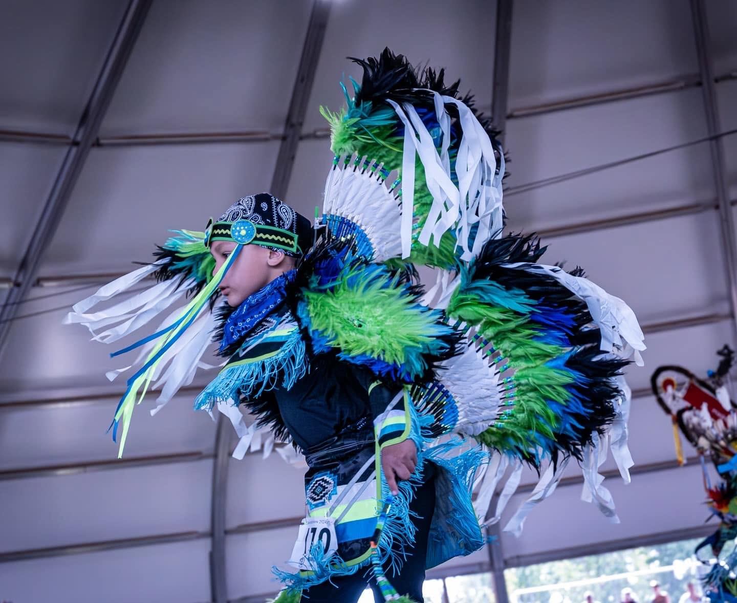 Dancer in bright green and blue feather regalia performs indoors on a stage.