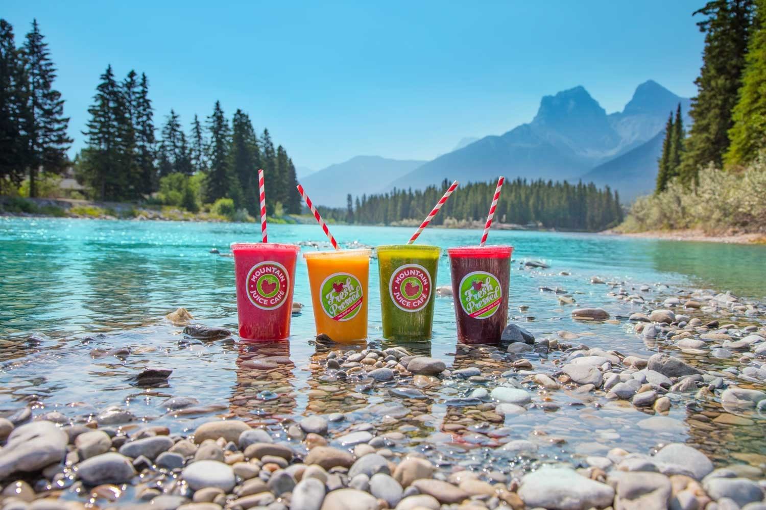 Four colorful drinks with striped straws on rocky riverbed, surrounded by water, trees, and mountains.