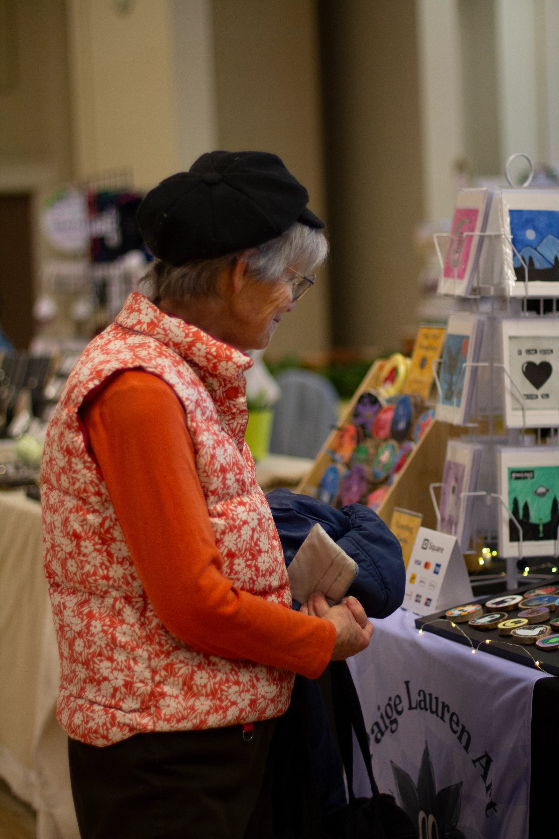 Shopper browsing handmade cards, prints, and small gifts at a vendor table at an indoor market.