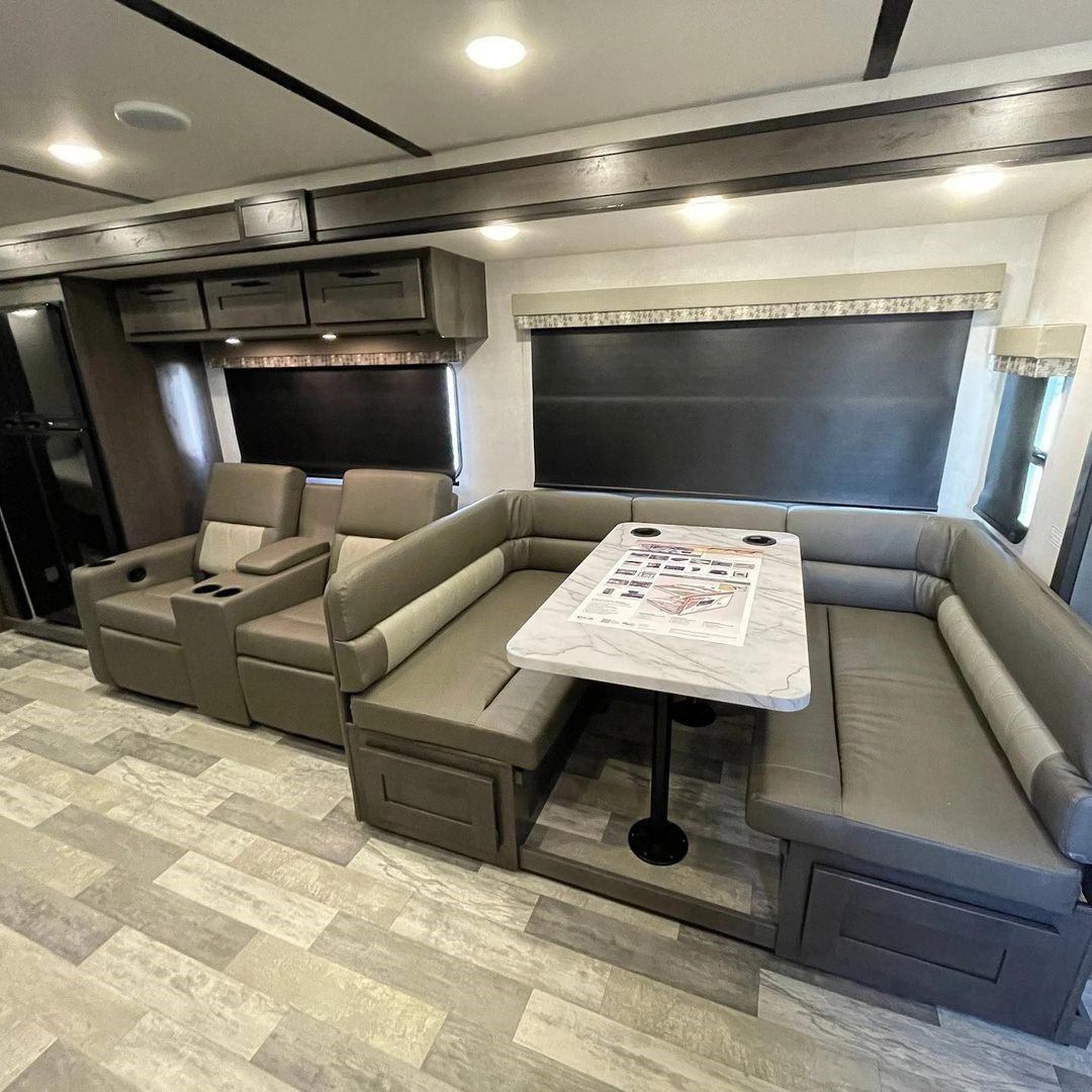 RV interior with U-shaped dinette, recliners, large windows, and wood-style flooring.