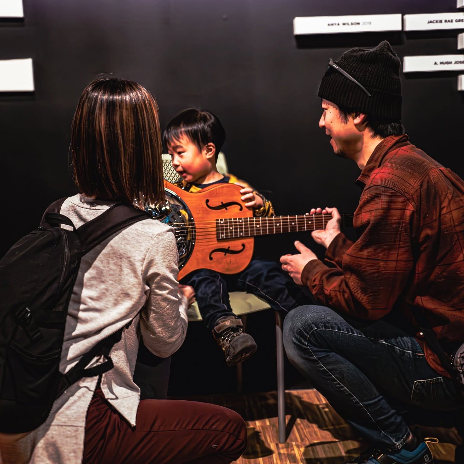 Child holds a guitar while two people sit nearby.
