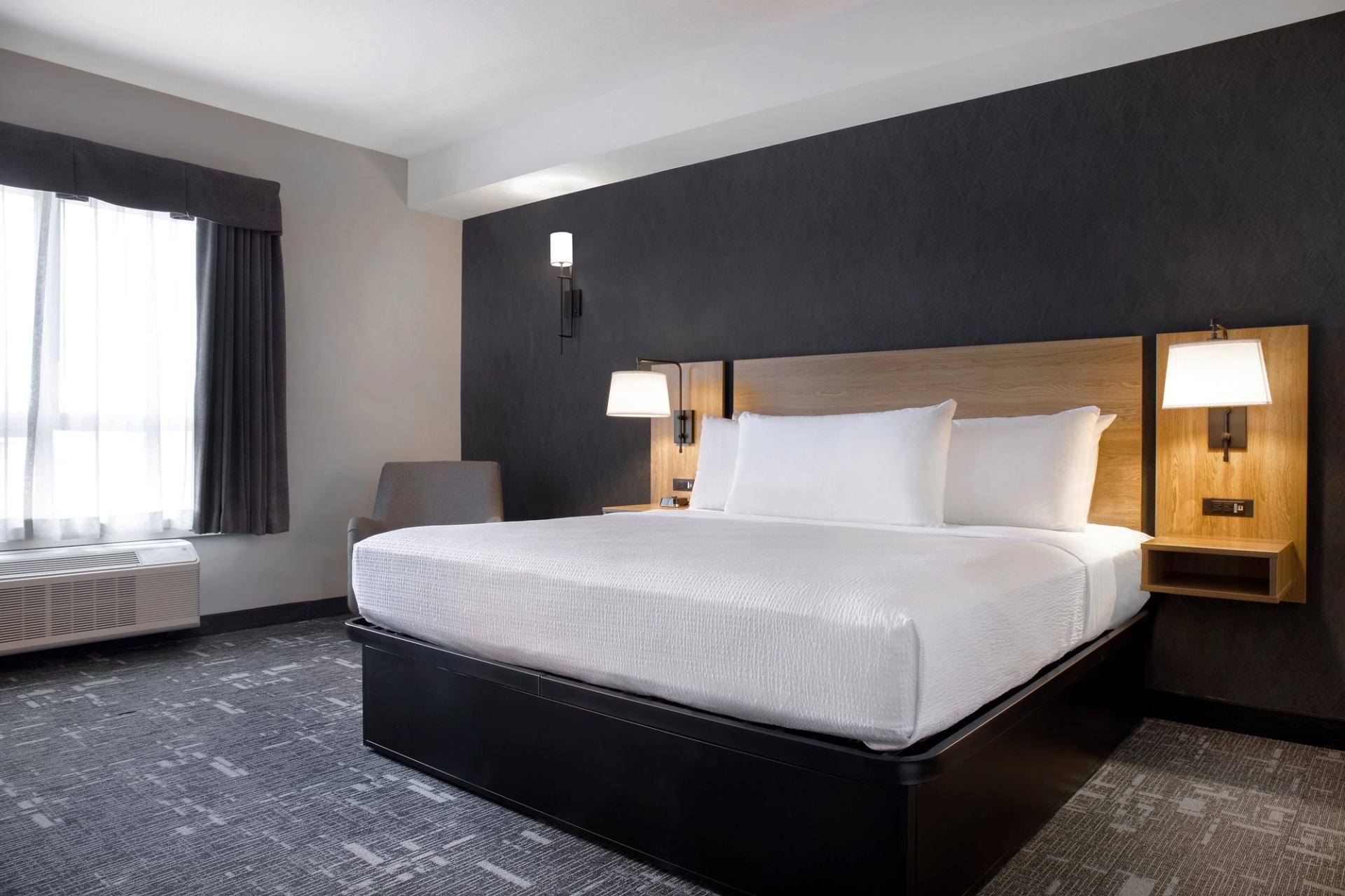 Hotel room with large bed, grey accent wall, lamps and window with curtains.