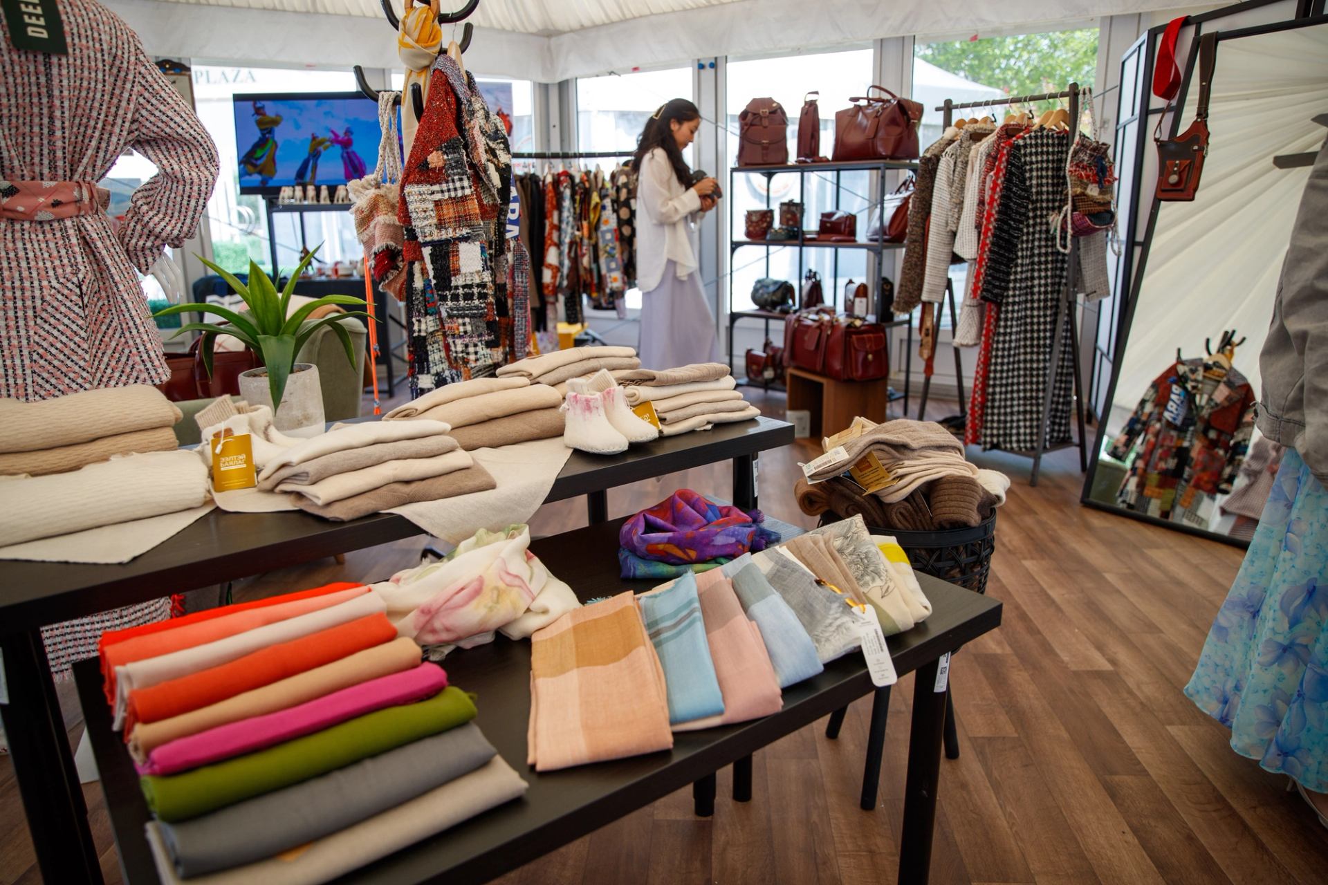 An indoor market stall displaying folded textiles, clothing on racks, and leather bags, with a woman browsing.