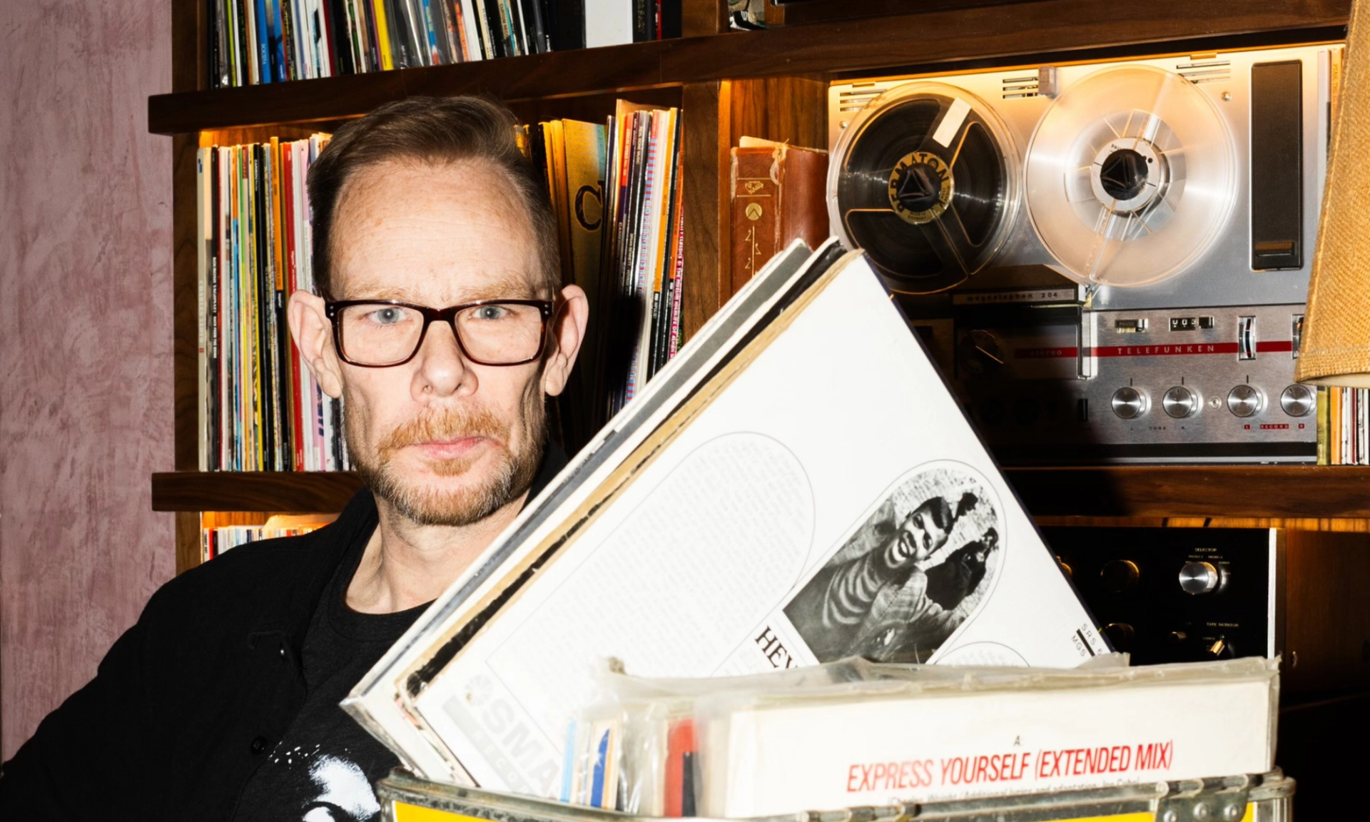 Person with glasses stands by shelves of vinyl records and reel-to-reel player.