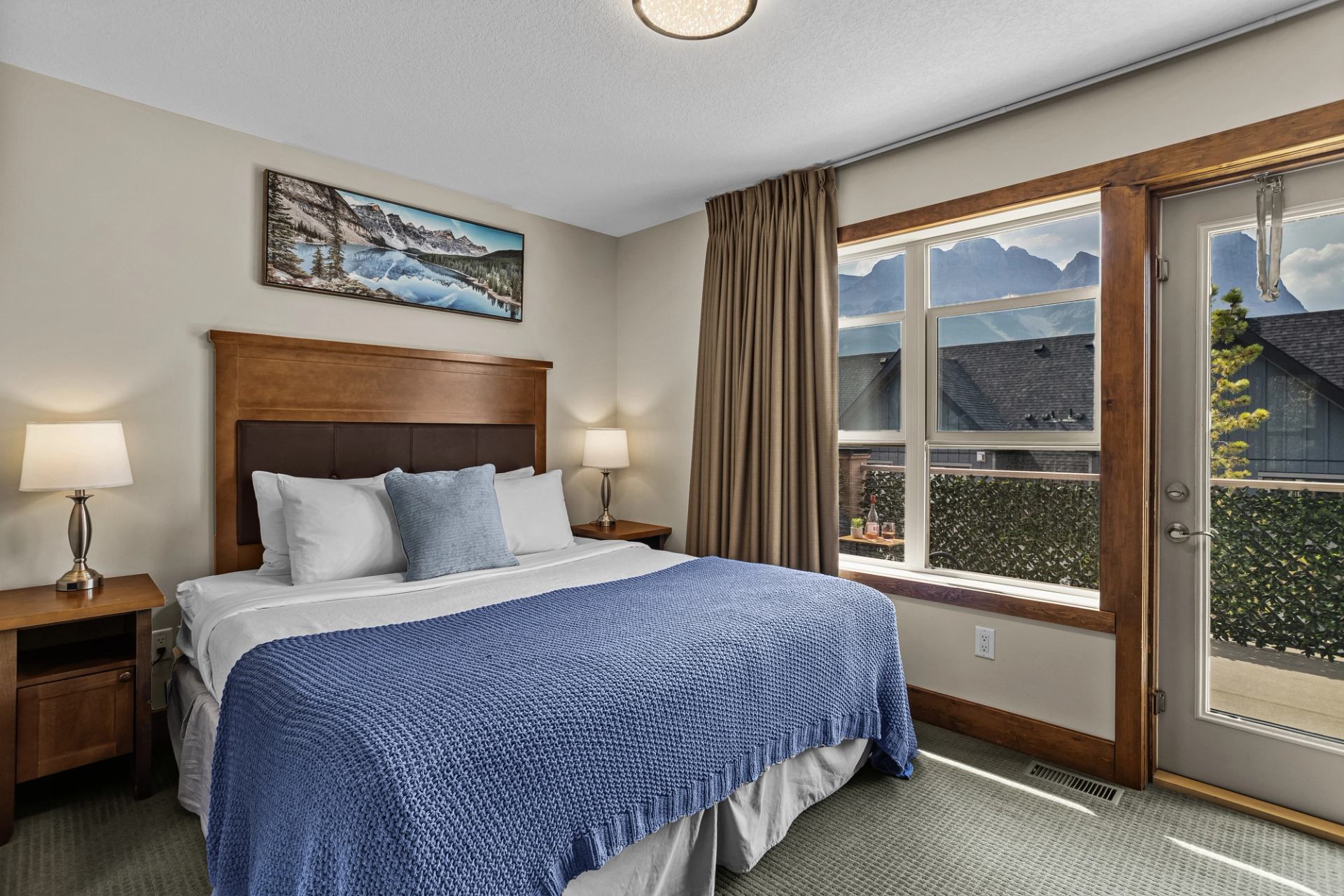 Bedroom with blue-covered bed, wooden furniture and windows overlooking mountains.