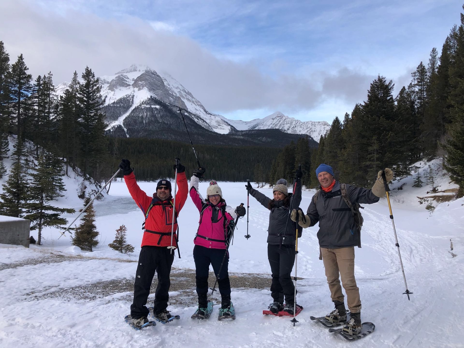  Four hikers on snowshoes posing with raised poles near a frozen lake and mountains.