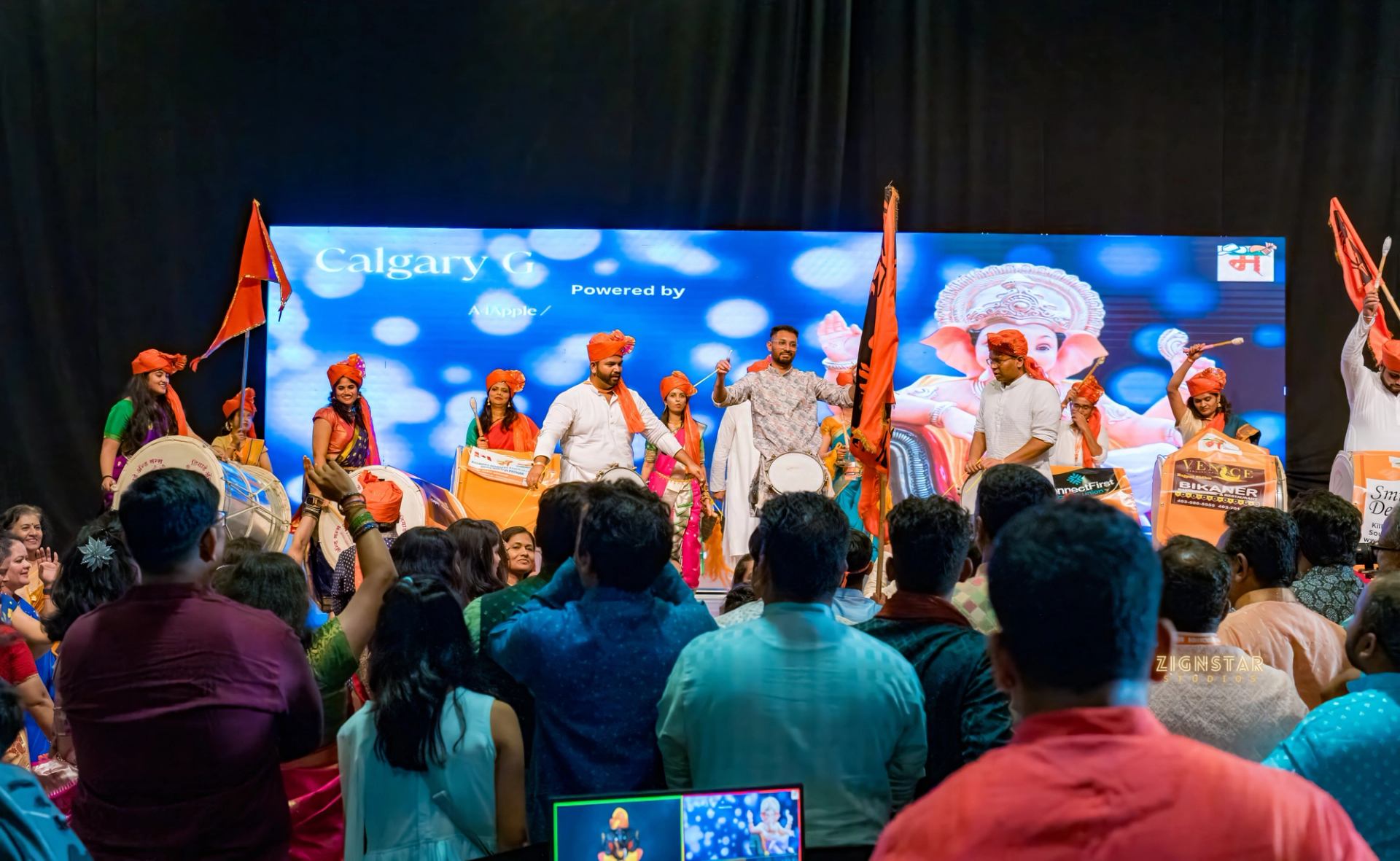 Drummers in orange headwear perform onstage before a standing crowd.