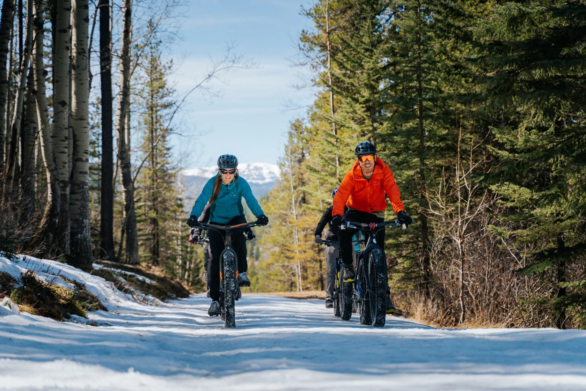 Mountain bikers ride on a snowy trail through a forest with scattered trees and brush.