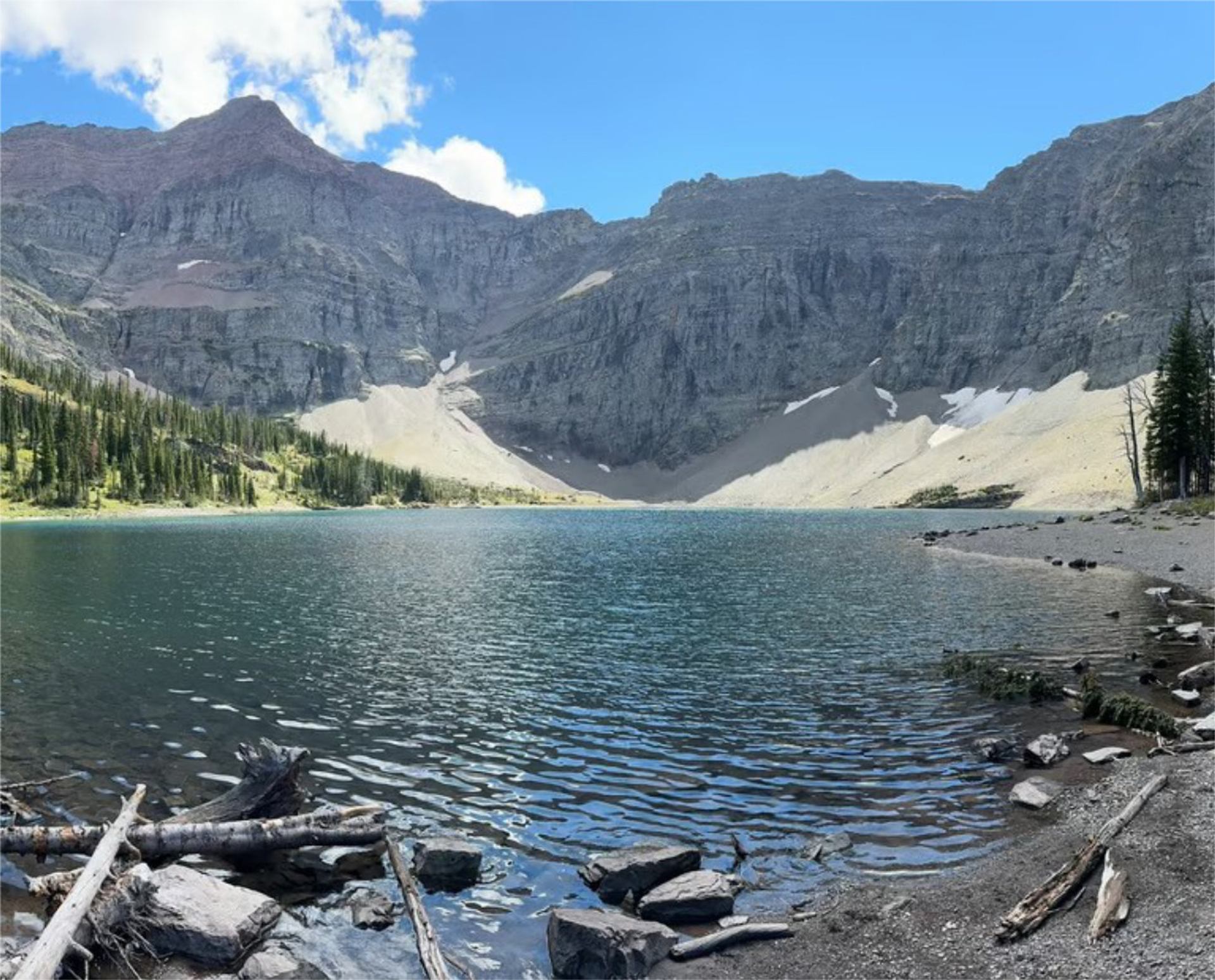 Mountain lake with rocky shoreline, evergreen trees and steep grey cliffs under blue sky.