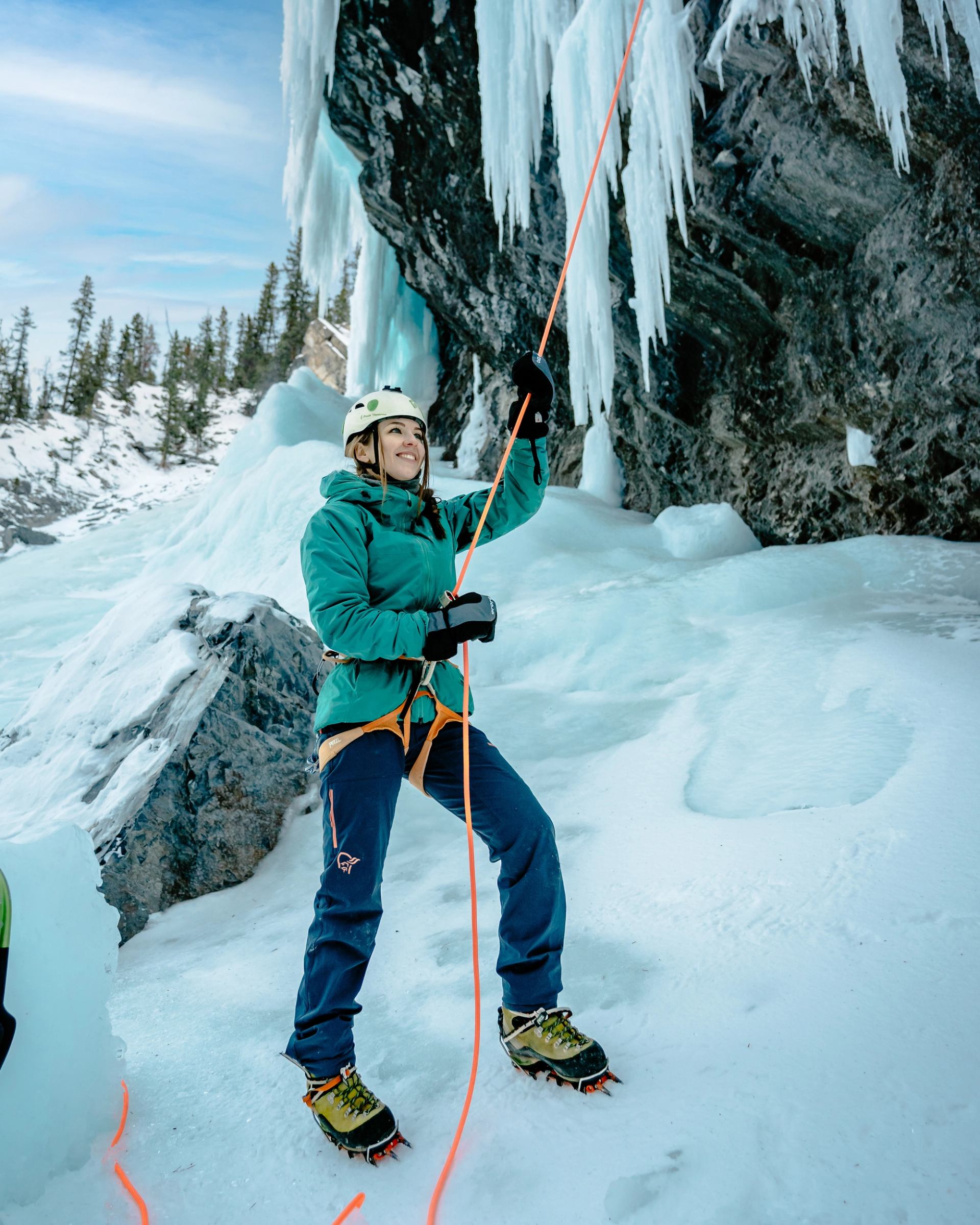 Woman in ice climbing gear and crampons holds a rope, standing below a large cliff covered in icicles.