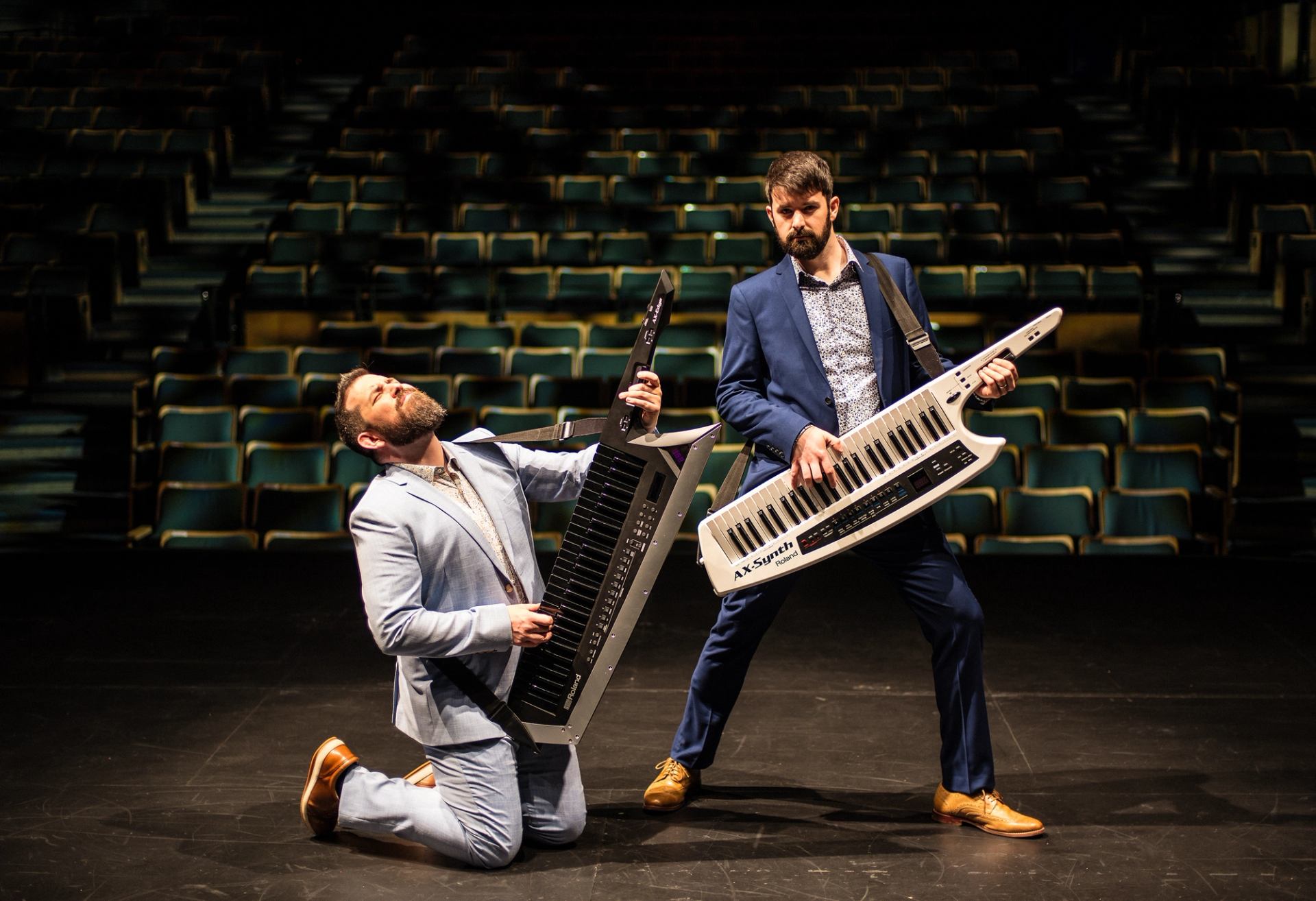 Two suited performers play keytars on a theatre stage before empty green seats.
