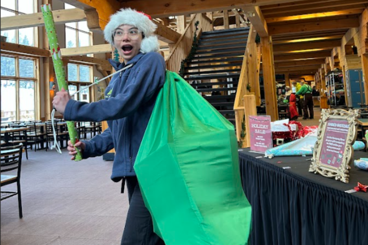 Person carries a large green gift bag and wrapping paper inside a lodge decorated for holidays.