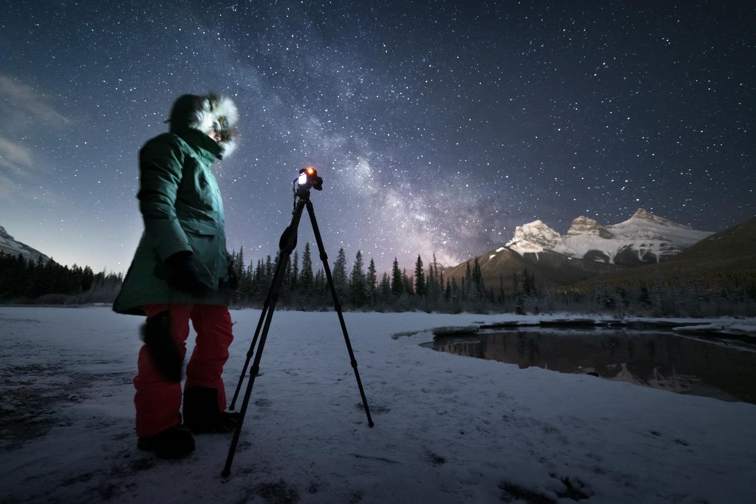 Person stands by a tripod camera on snowy ground under a star-filled sky and Milky Way.
