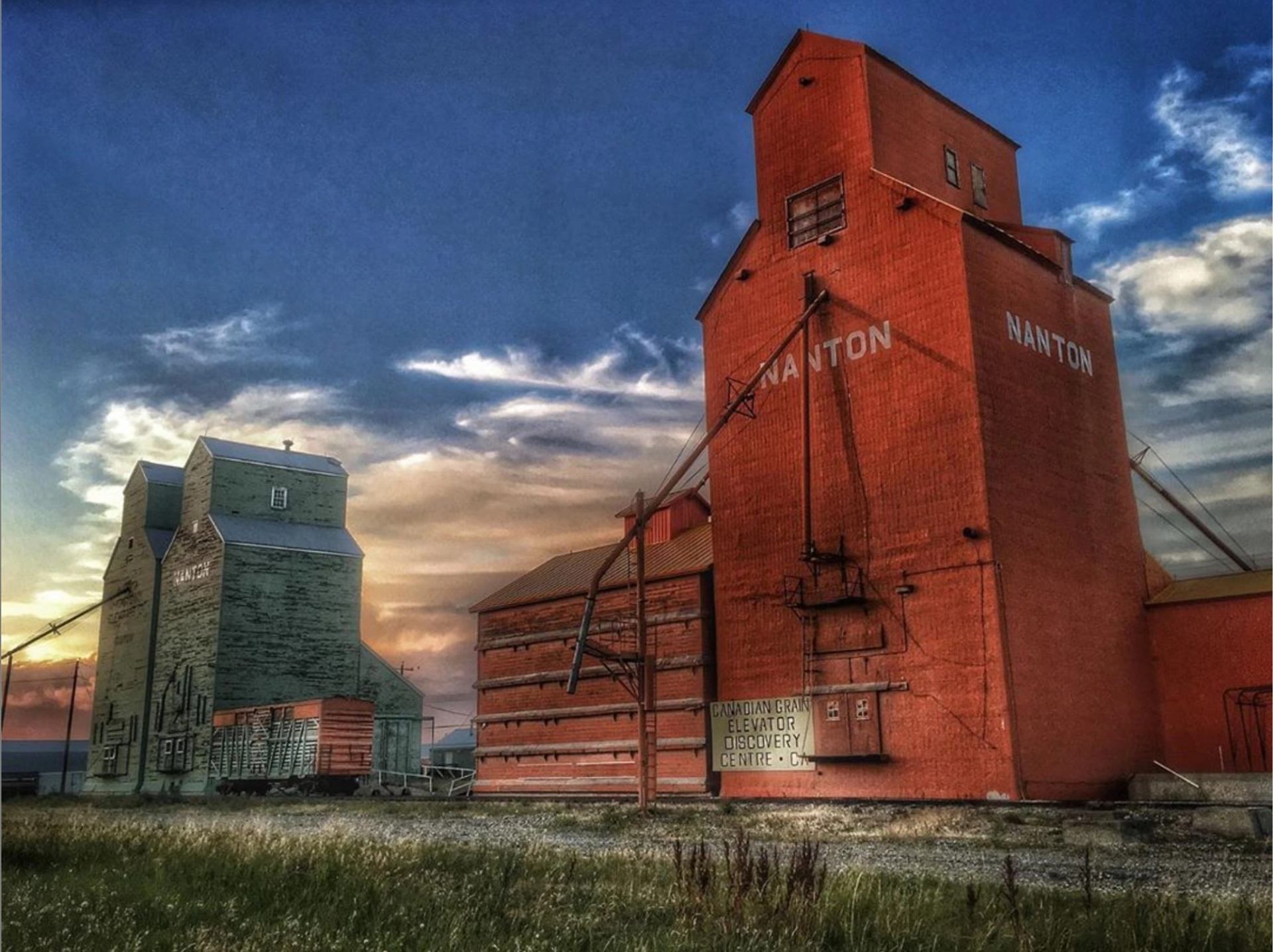 Red and green grain elevators marked “Nanton” under a colorful evening sky