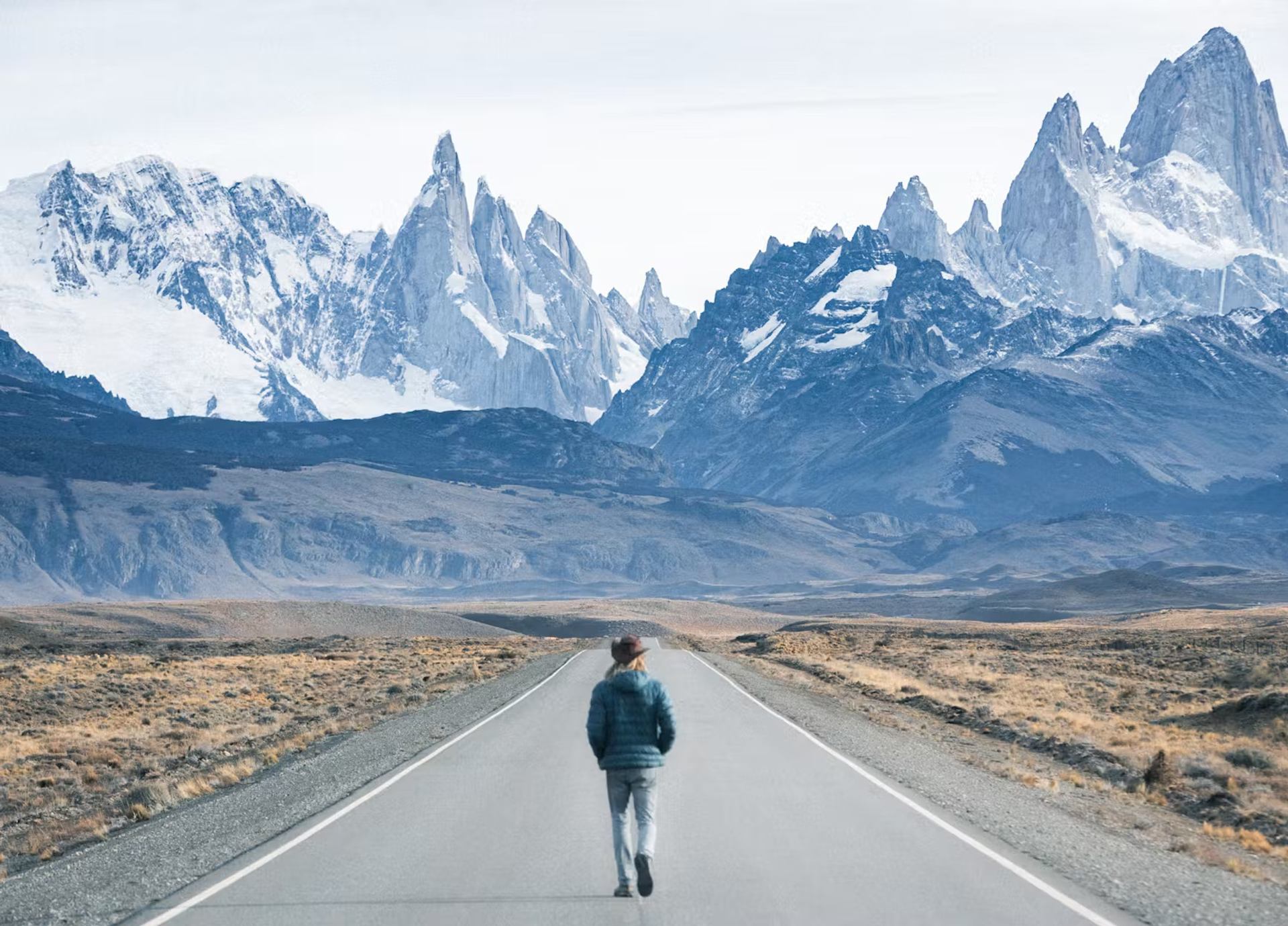 Person walking a long road toward jagged, snow-capped mountains in a vast, rugged landscape.