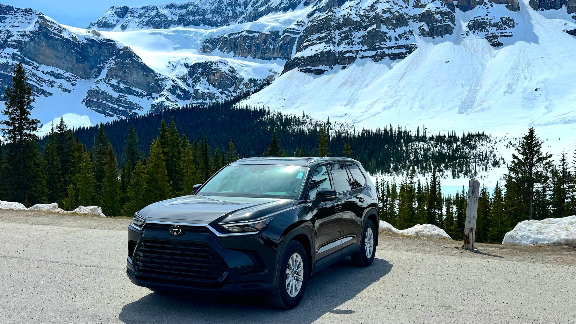 Black Toyota SUV parked on roadside with snow-covered mountains and evergreen forest behind.