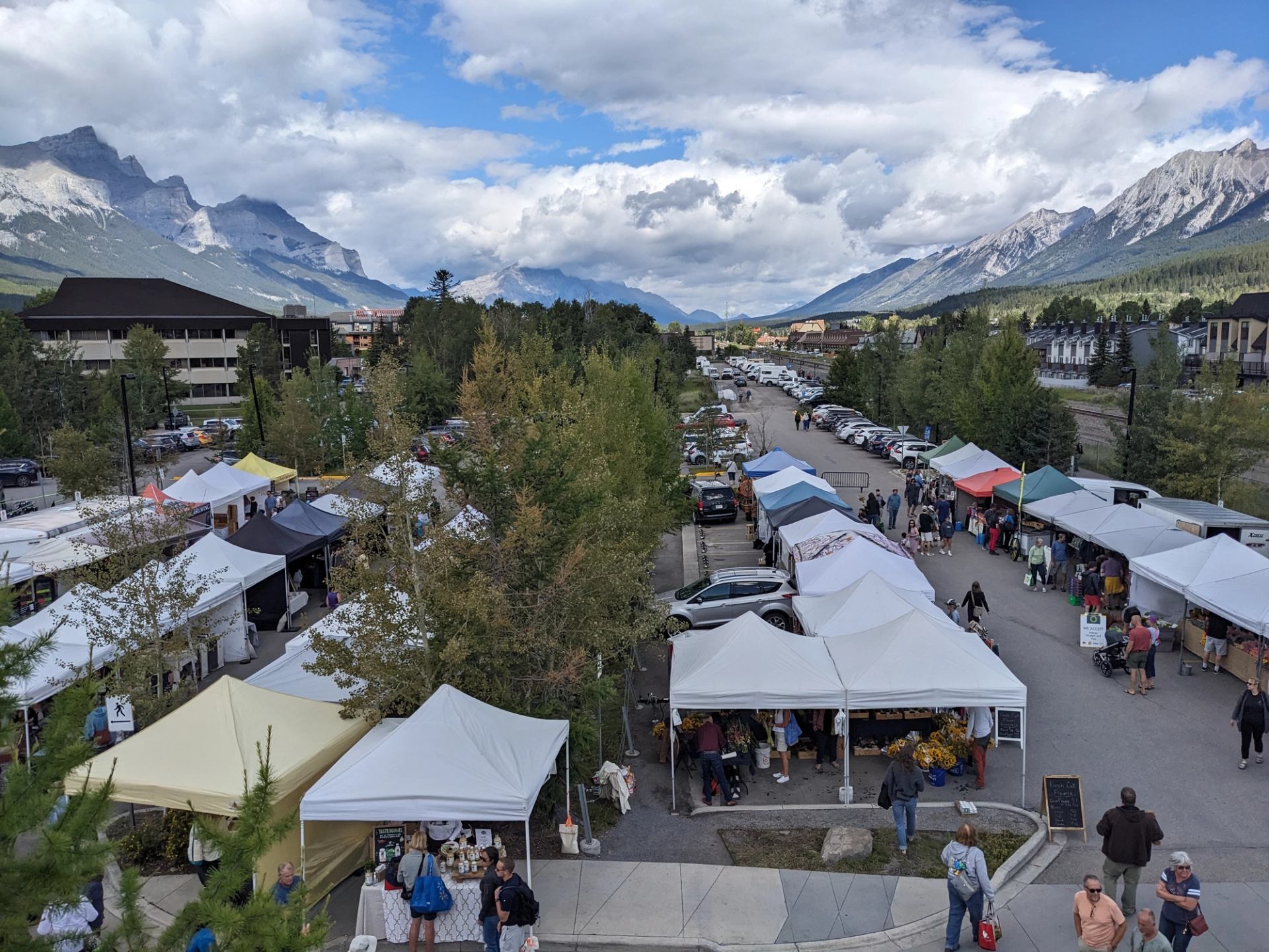 Market tents and parked cars at Canmore Mountain Market with mountain views and cloudy sky.