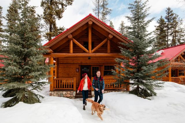 Couple and dog walking in the snow outside a log cabin at Baker Creek Mountain Resort