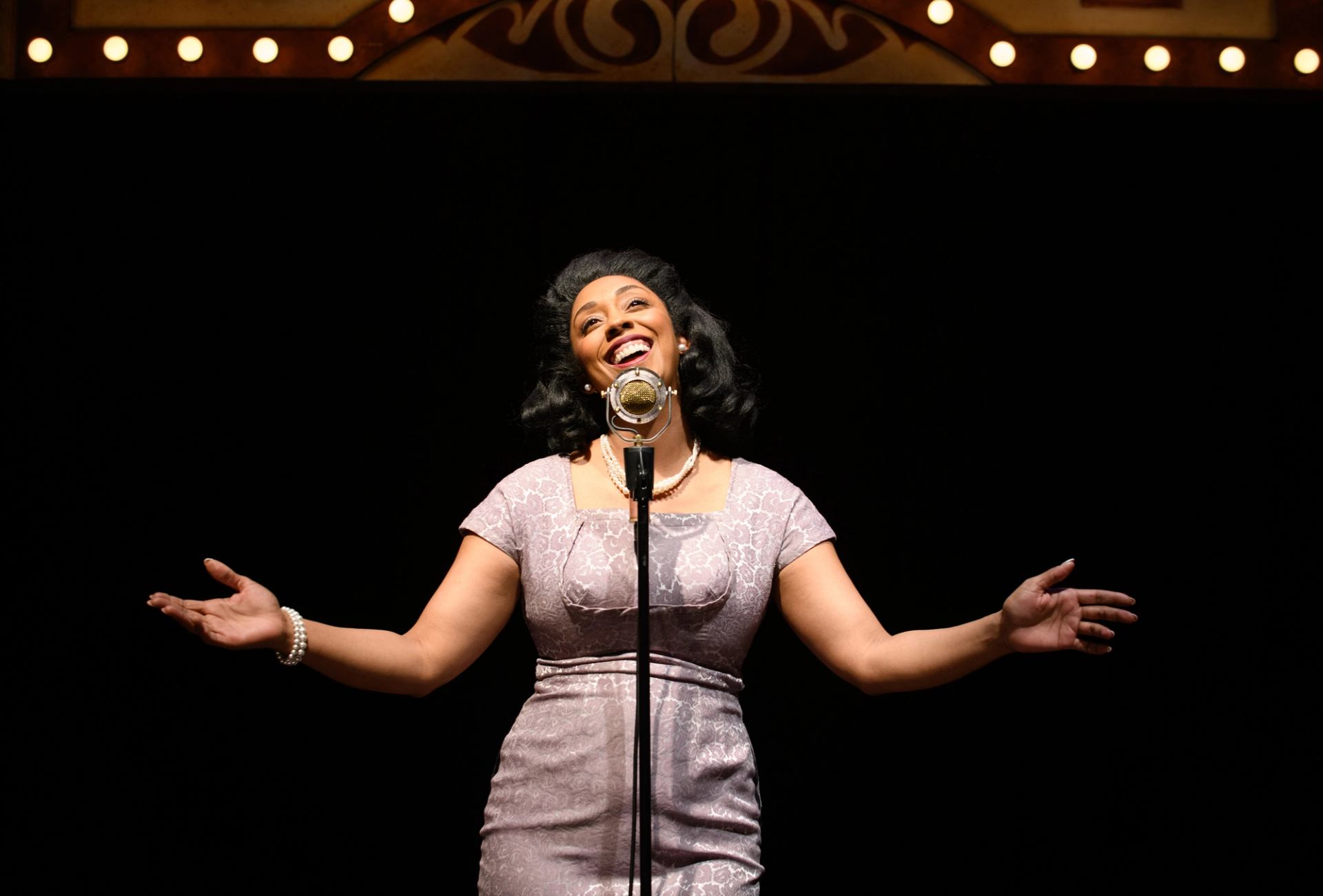 Singer in vintage dress performs at old-fashioned microphone under theatre lights.