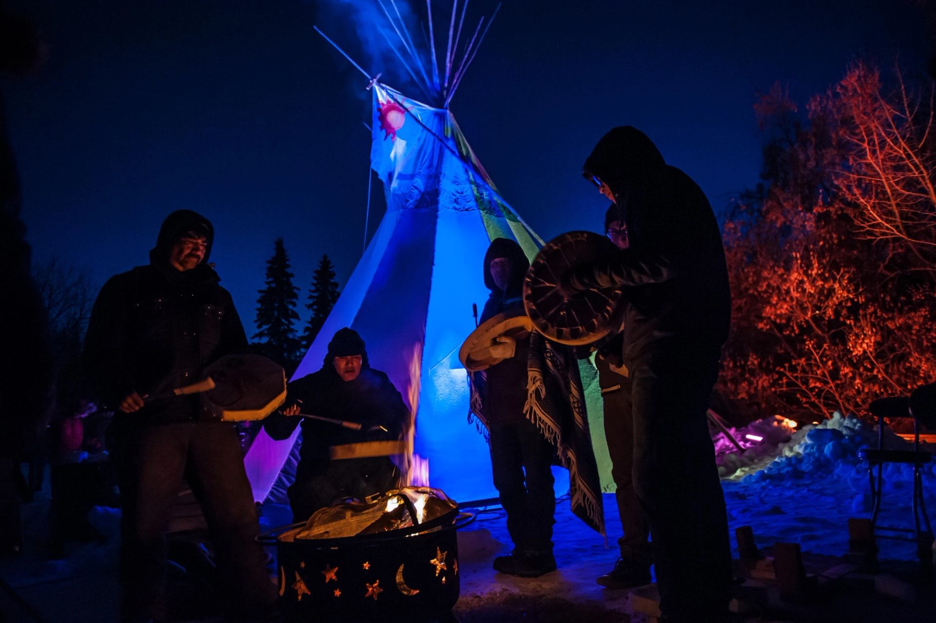 Group playing drums near a glowing tipi and fire pit at Flying Canoë Volant festival.