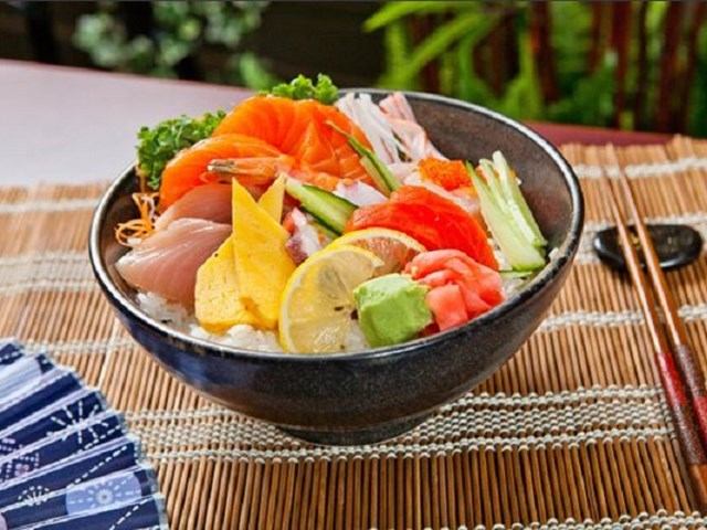 Bowl of sashimi with salmon, vegetables, and lemon on a bamboo placemat.