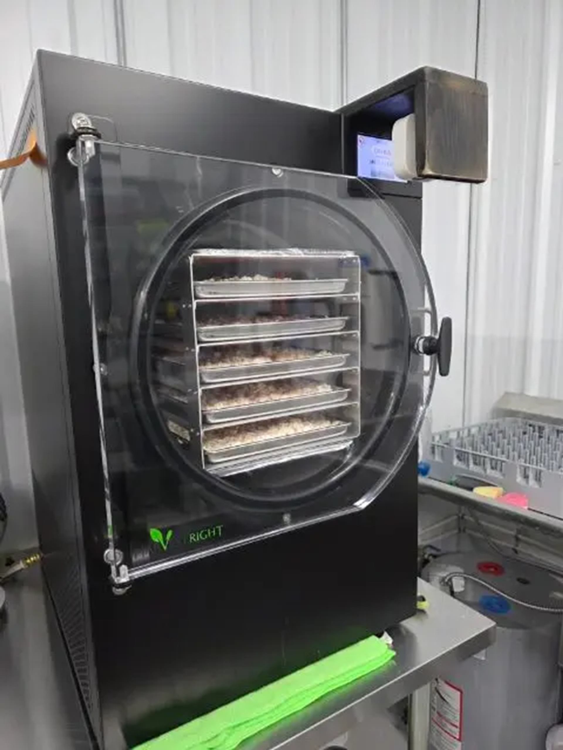 Black industrial freeze dryer with transparent door and food trays inside on metal table.