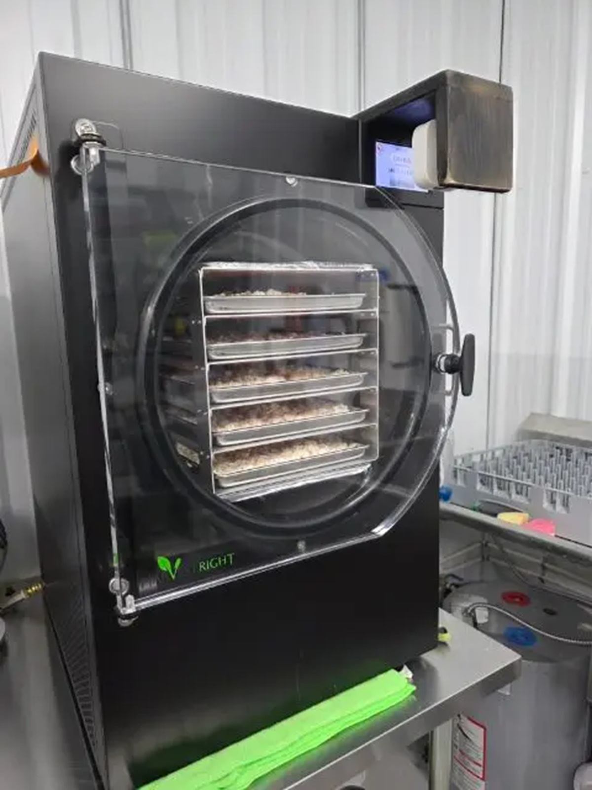 Black industrial freeze dryer with transparent door and food trays inside on metal table.