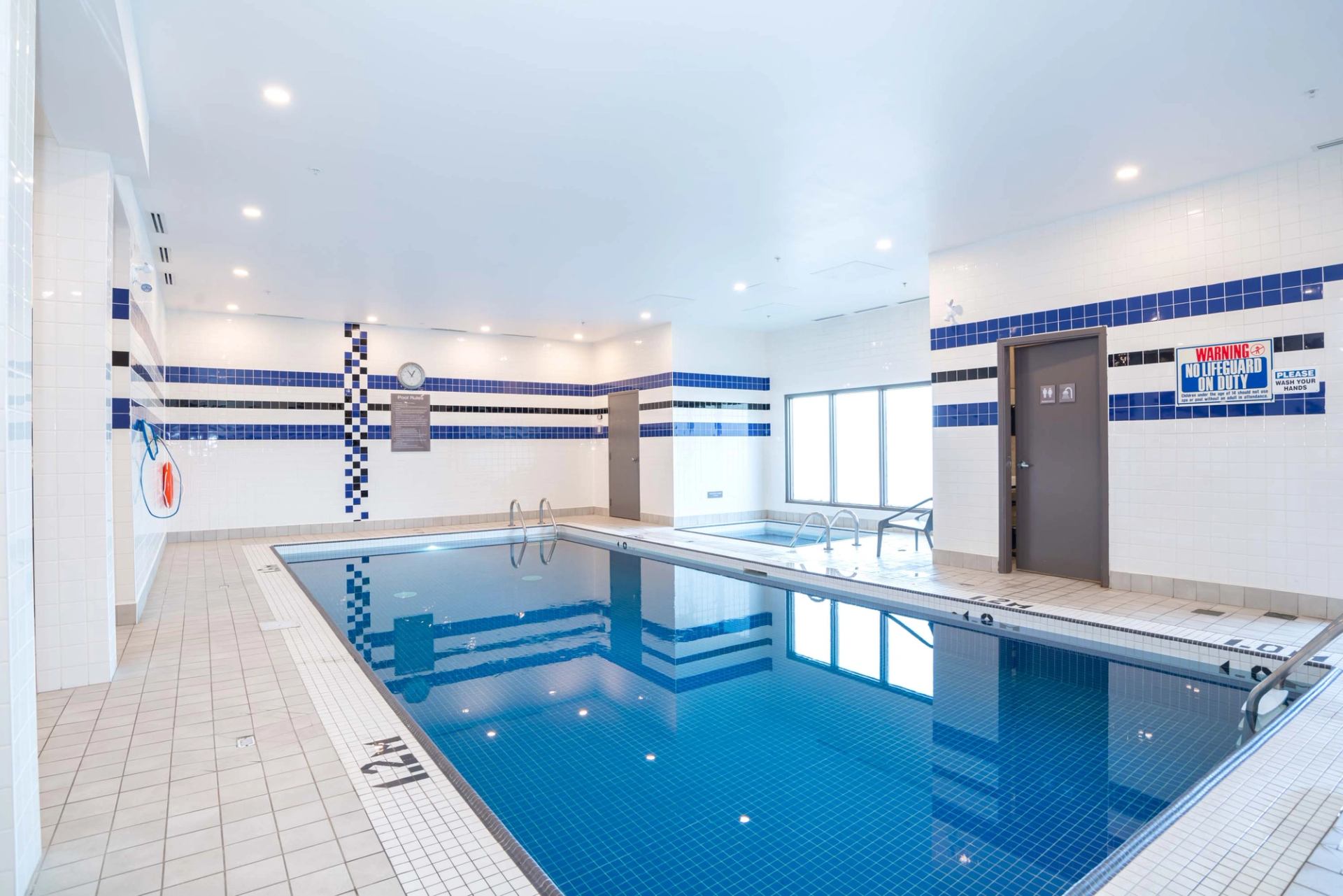 Indoor swimming pool area with blue tile, hot tub, safety signs and pool ladders.