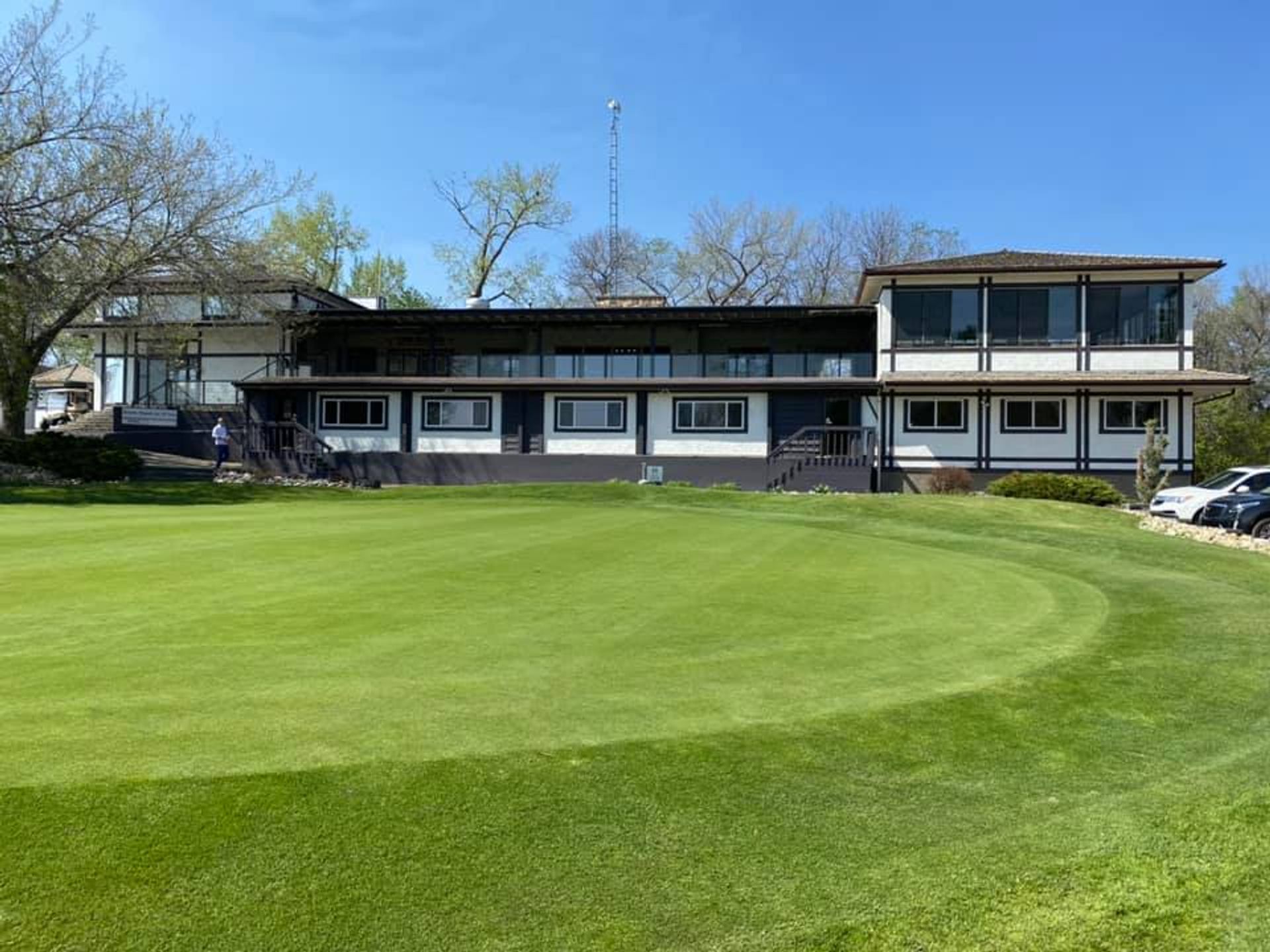 Clubhouse-style building behind manicured golf green, with trees and parked cars at sides.